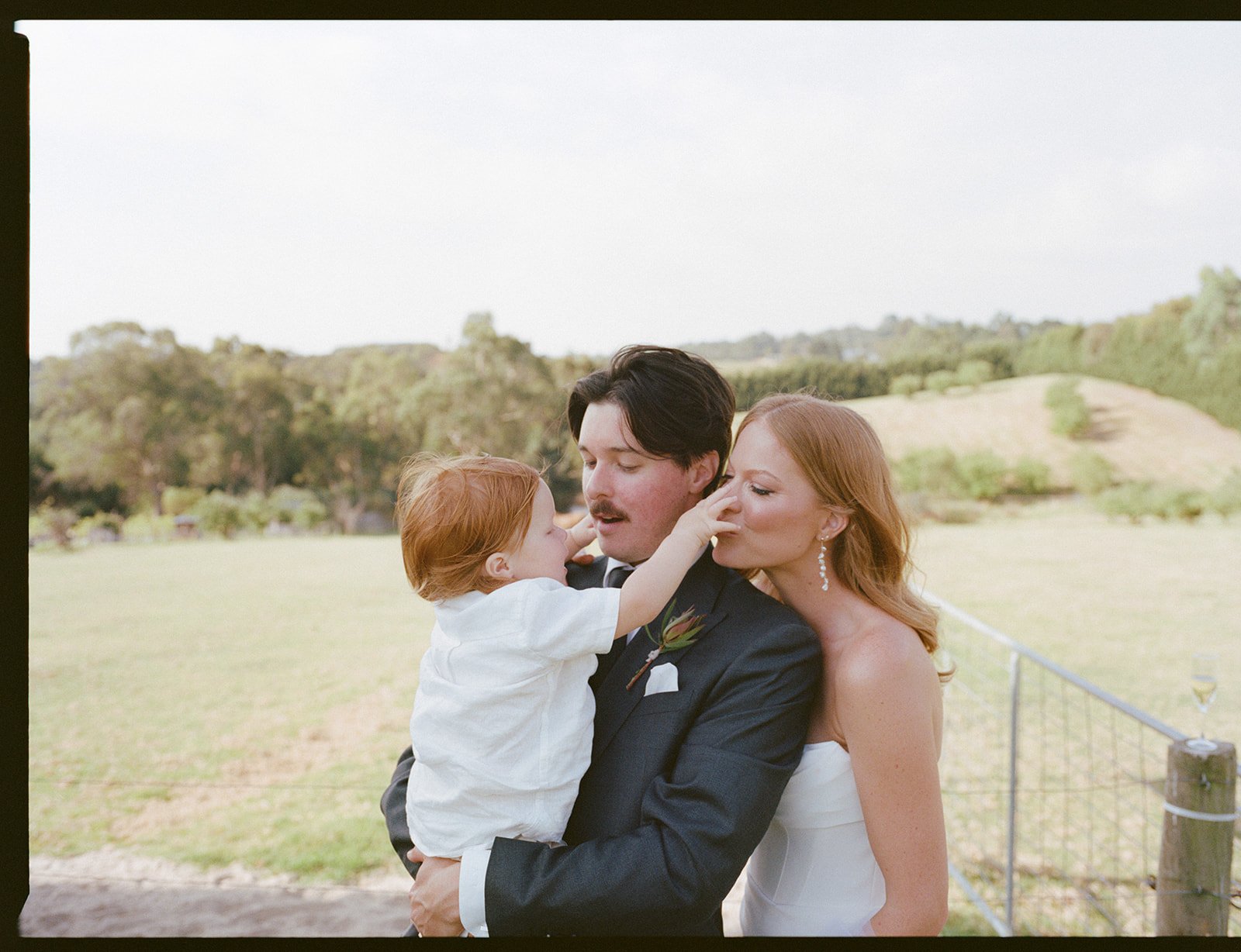 A family at an outdoor wedding, with a bride, groom, and a young child, where the child is reaching towards the grooms face.