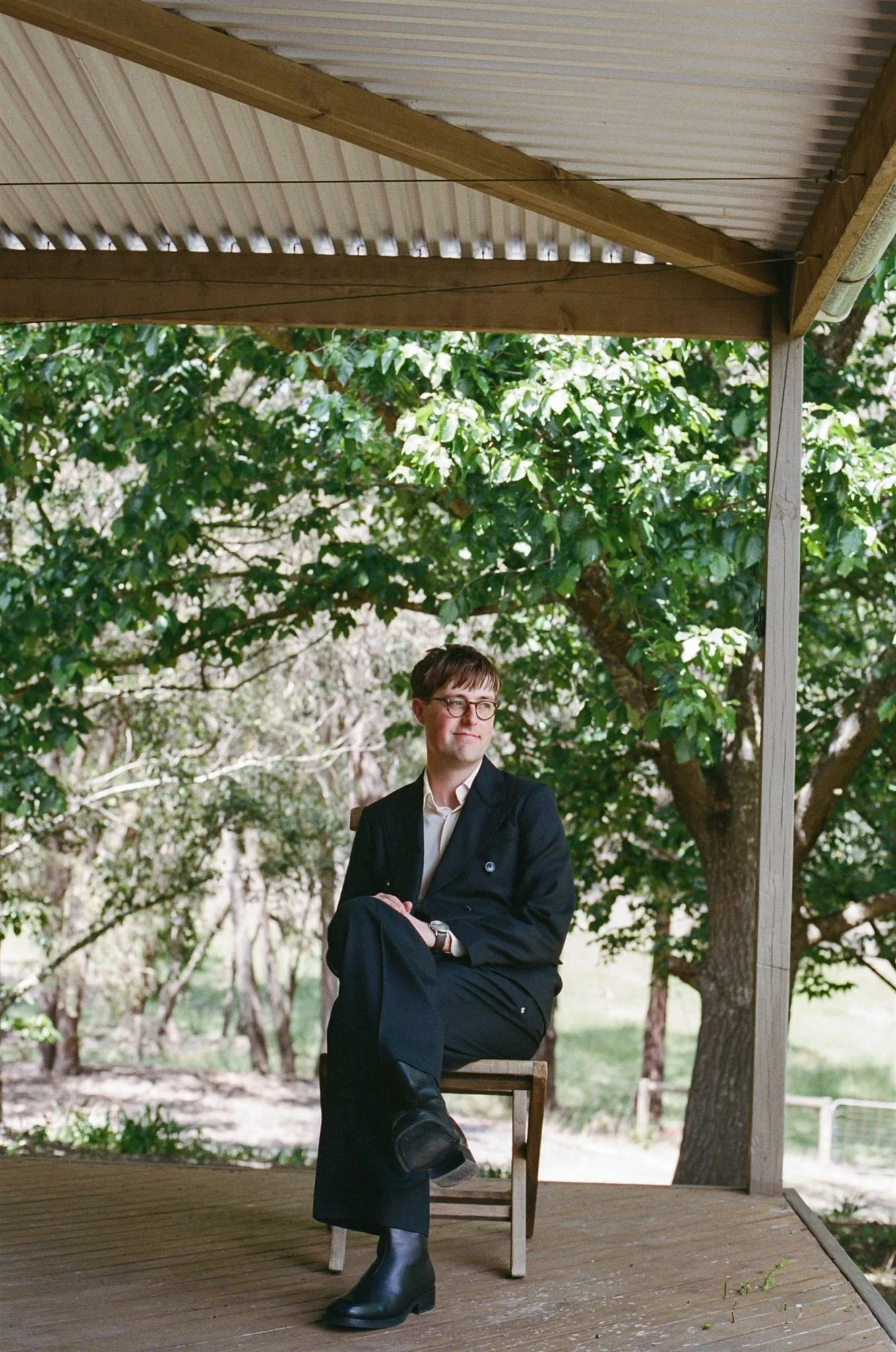 A groom wearing glasses and a black suit sits on a wooden chair on a porch with a wooden roof, surrounded by green trees in the background.