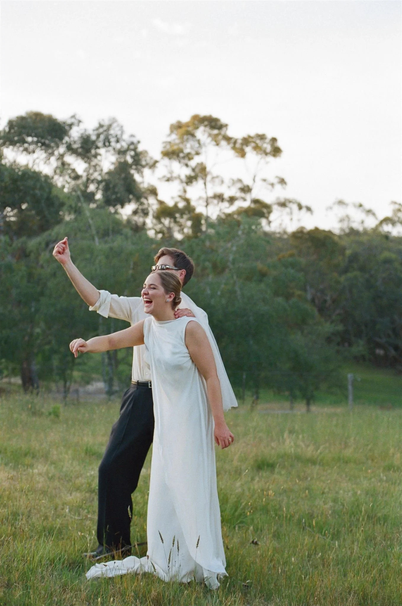 A bride and groom stand in a field with long grass they are smiling and waving at their friends in the distance. 