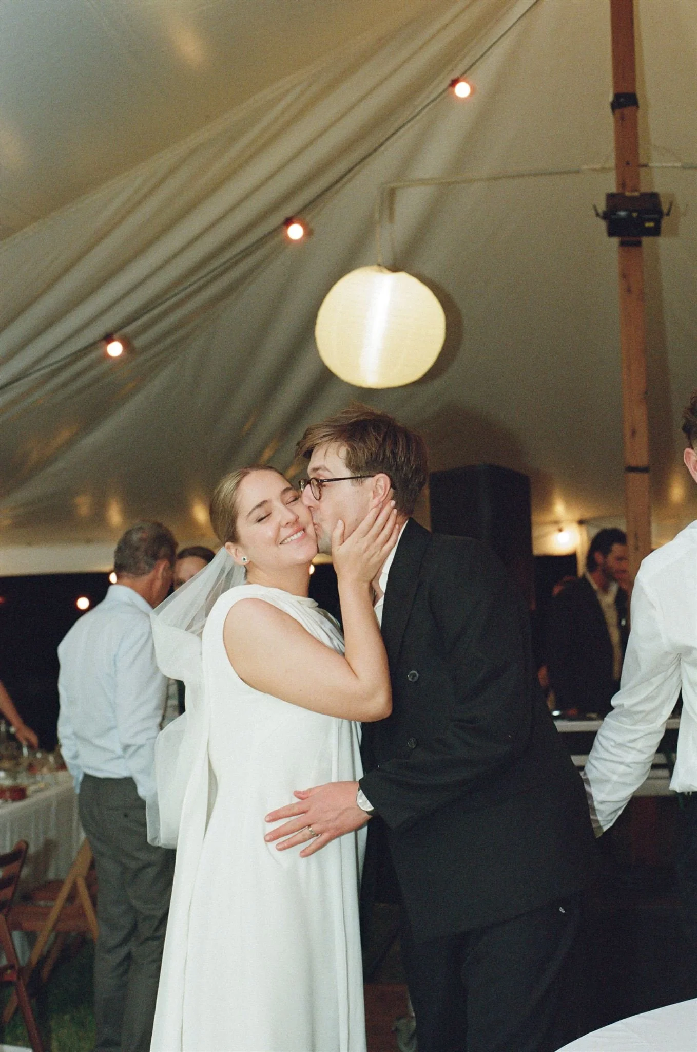 A groom kisses a brides cheek on the dance floor at their wedding. Lights glow behind them. 