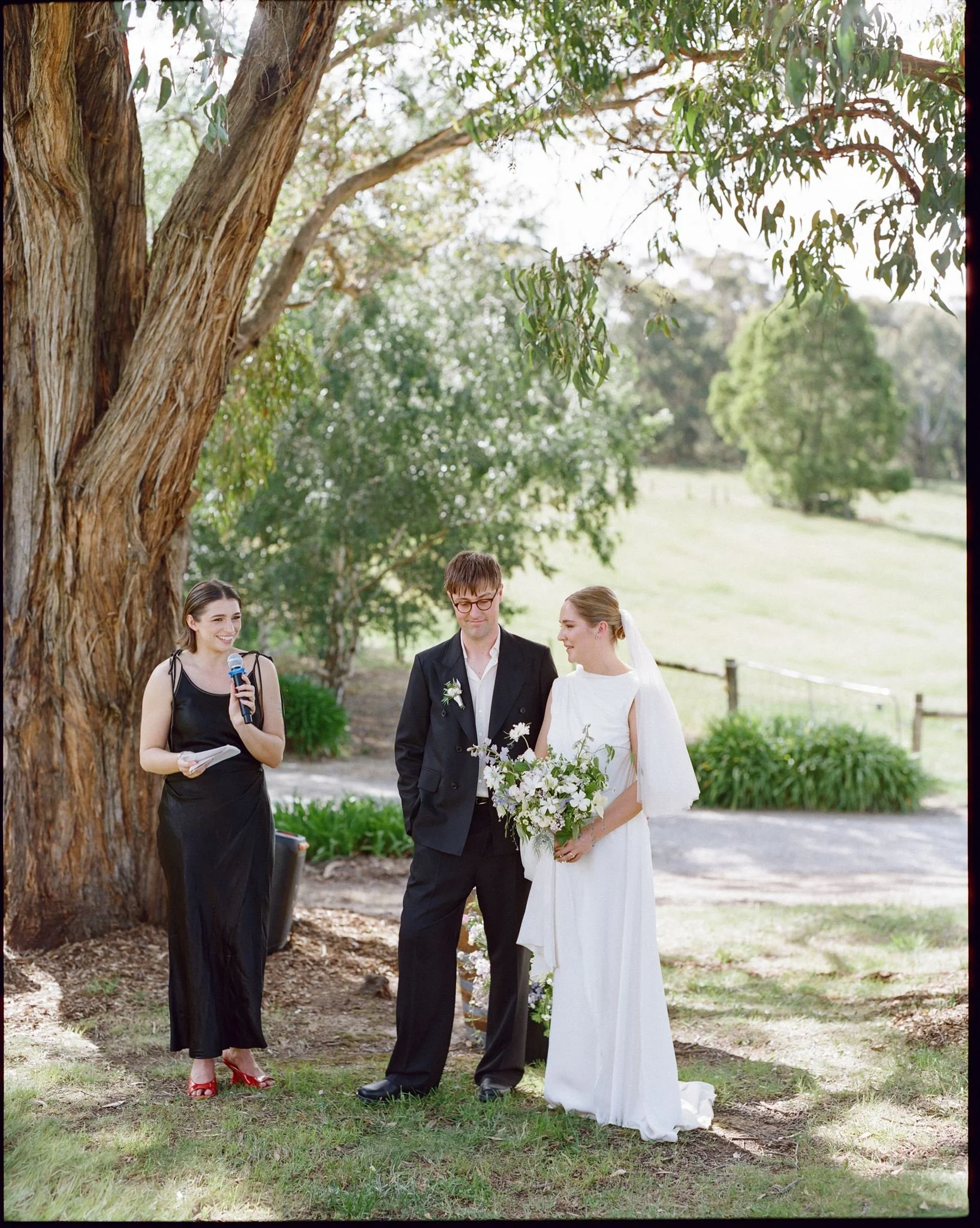 A bride and groom stand in front of a paddock during their wedding ceremony, their celebrant stands next to them. The bride holds a bouquet of blue, white, and green flowers.