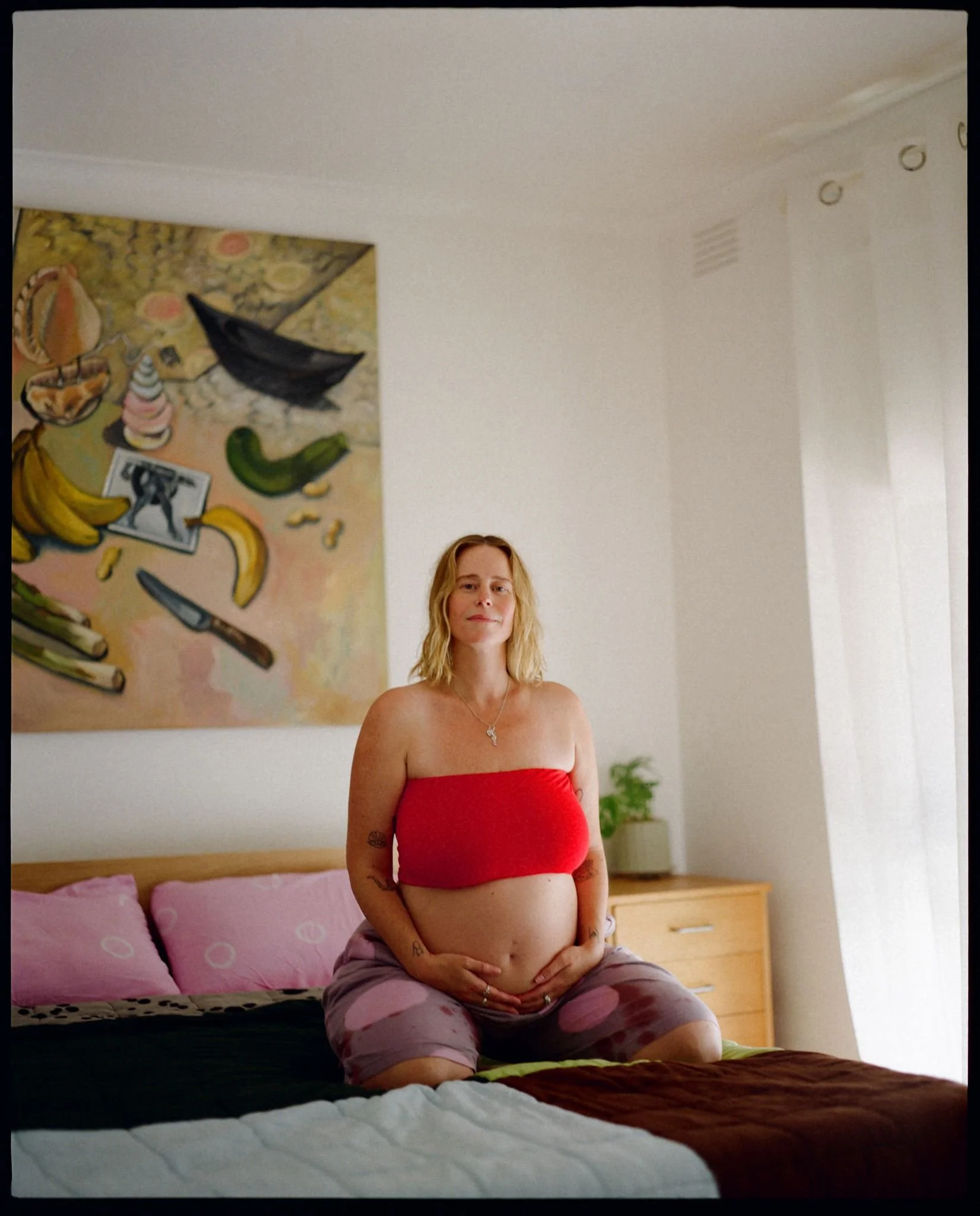 A pregnant women sits on her bed holding her tummy, light is streaming through the windows and there is a colourful artwork behind her.