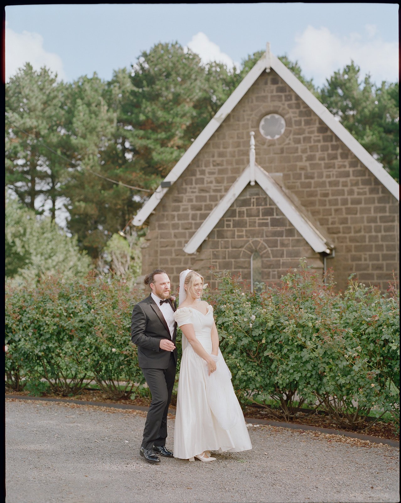 A bride and groom standing outdoors in front of a small stone church with large trees in the background. The groom is wearing a black tuxedo with a bow tie, and the bride is wearing a white wedding dress with puffed sleeves. The sky is partly cloudy.