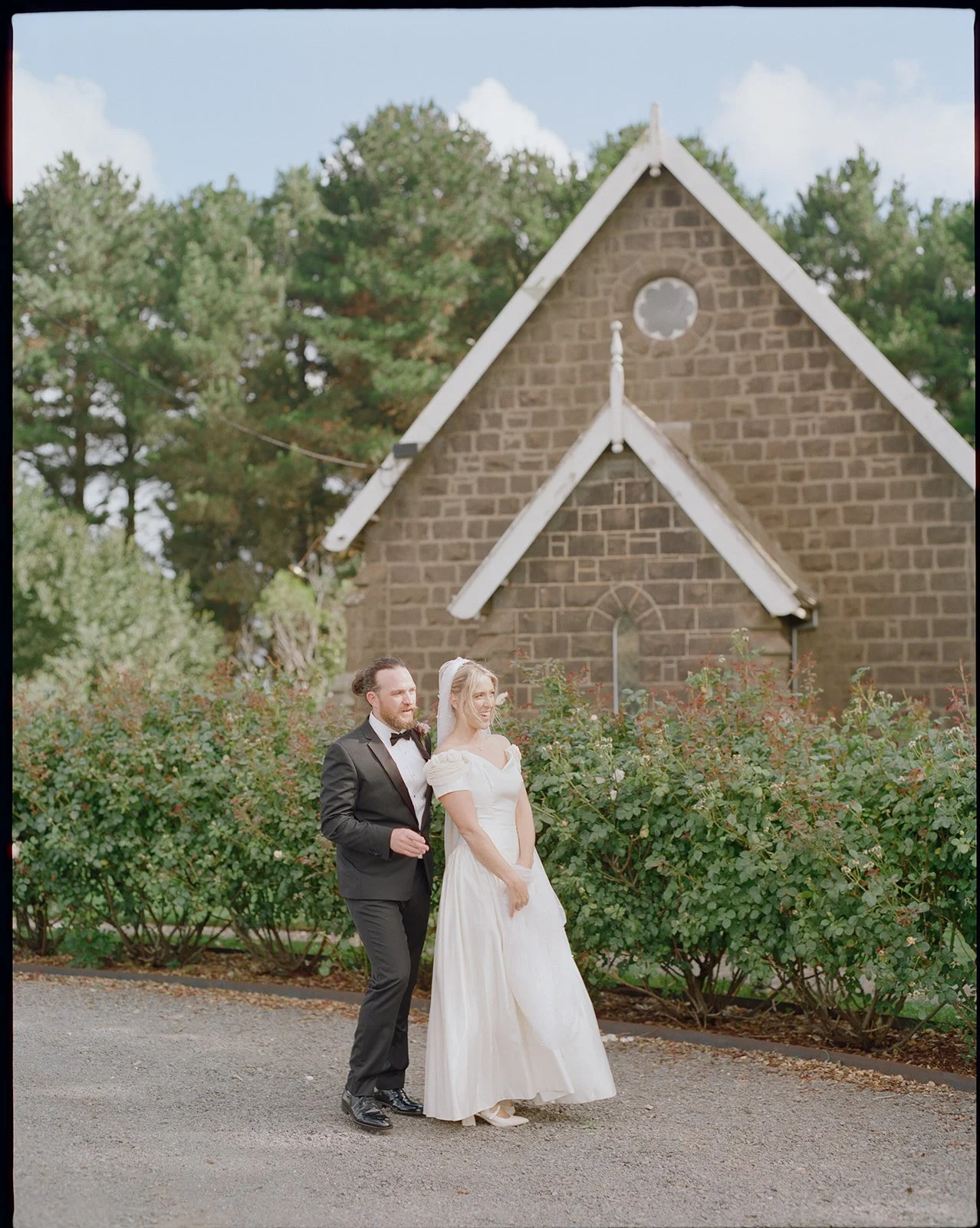 a bride and groom stand in front of a blue stone chapel, the a rose bushes in front, the bride wears a white vintage dress and the grooms wears a black tuxedo.