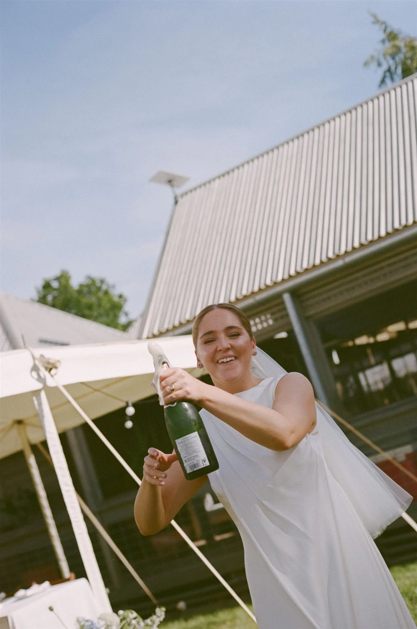 A bride pops a bottle of champagne and smiles, she is wearing a white sleeveless wedding dress and short veil.