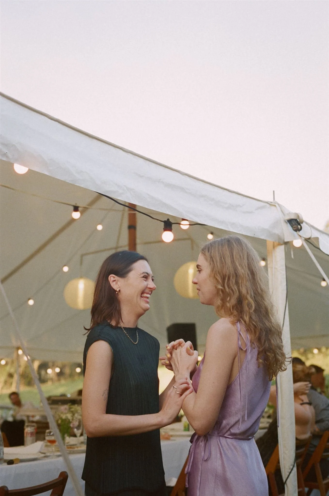 Two wedding guests are standing together chatting and smiling. one is wearing a black dress and the other a lilac silk dress. You can see fairy lights in the background. 
