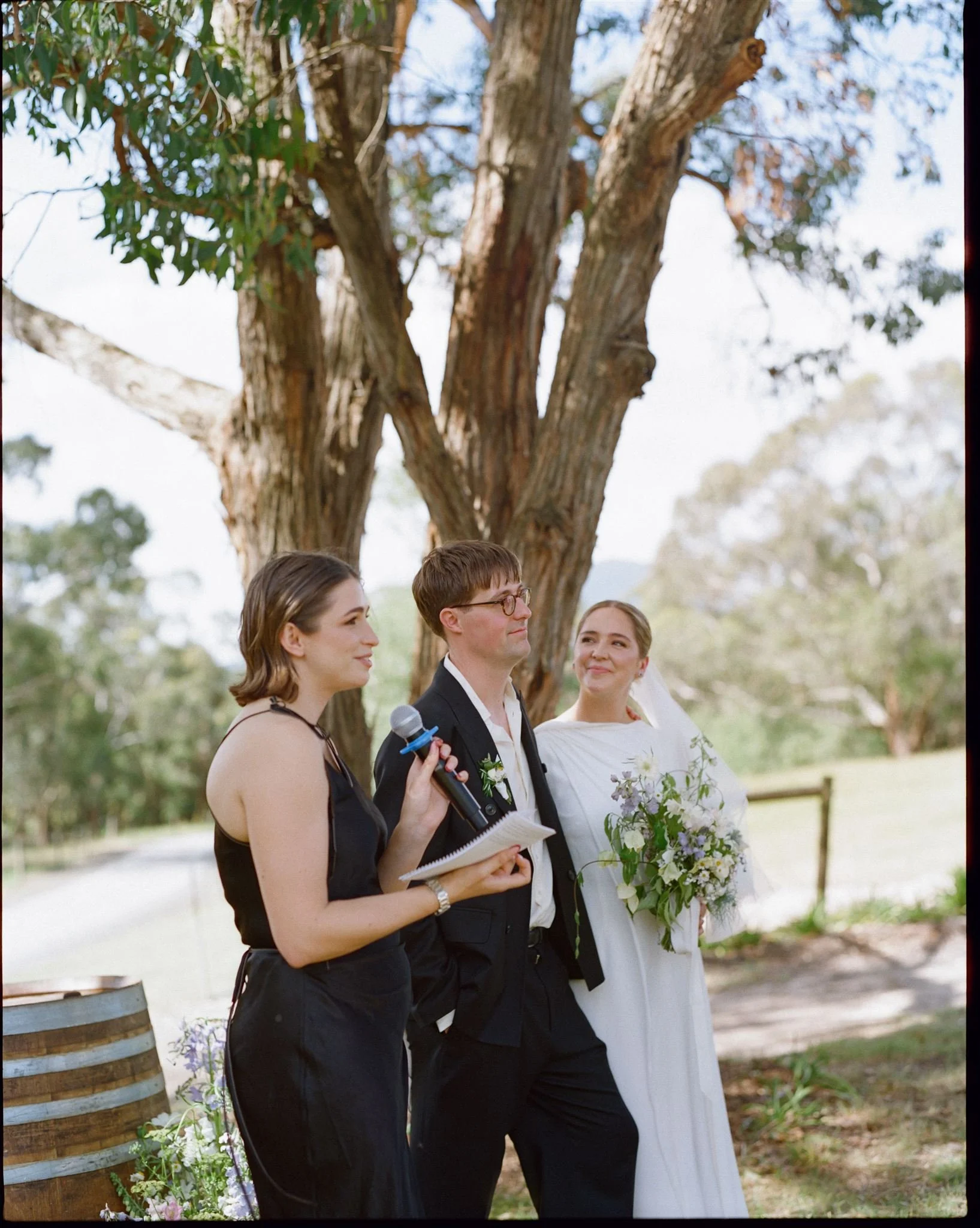 a bride and groom stand in front of a tree during their ceremony, their friend is hosting their ceremony, the bride looks lovingly at her groom.