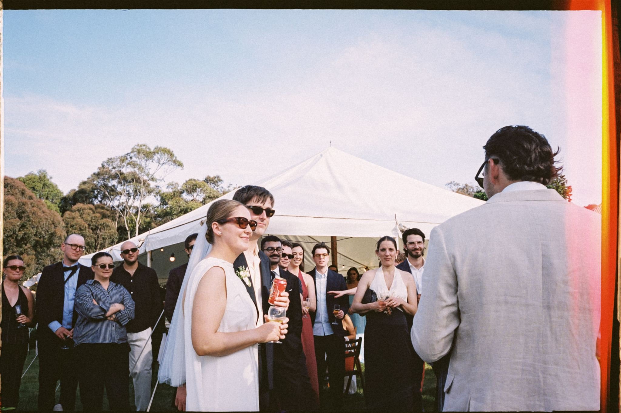Group of people at outdoor wedding reception under a white tent, with a clear sky and trees in the background, as a man in a light gray suit addresses the guests.
