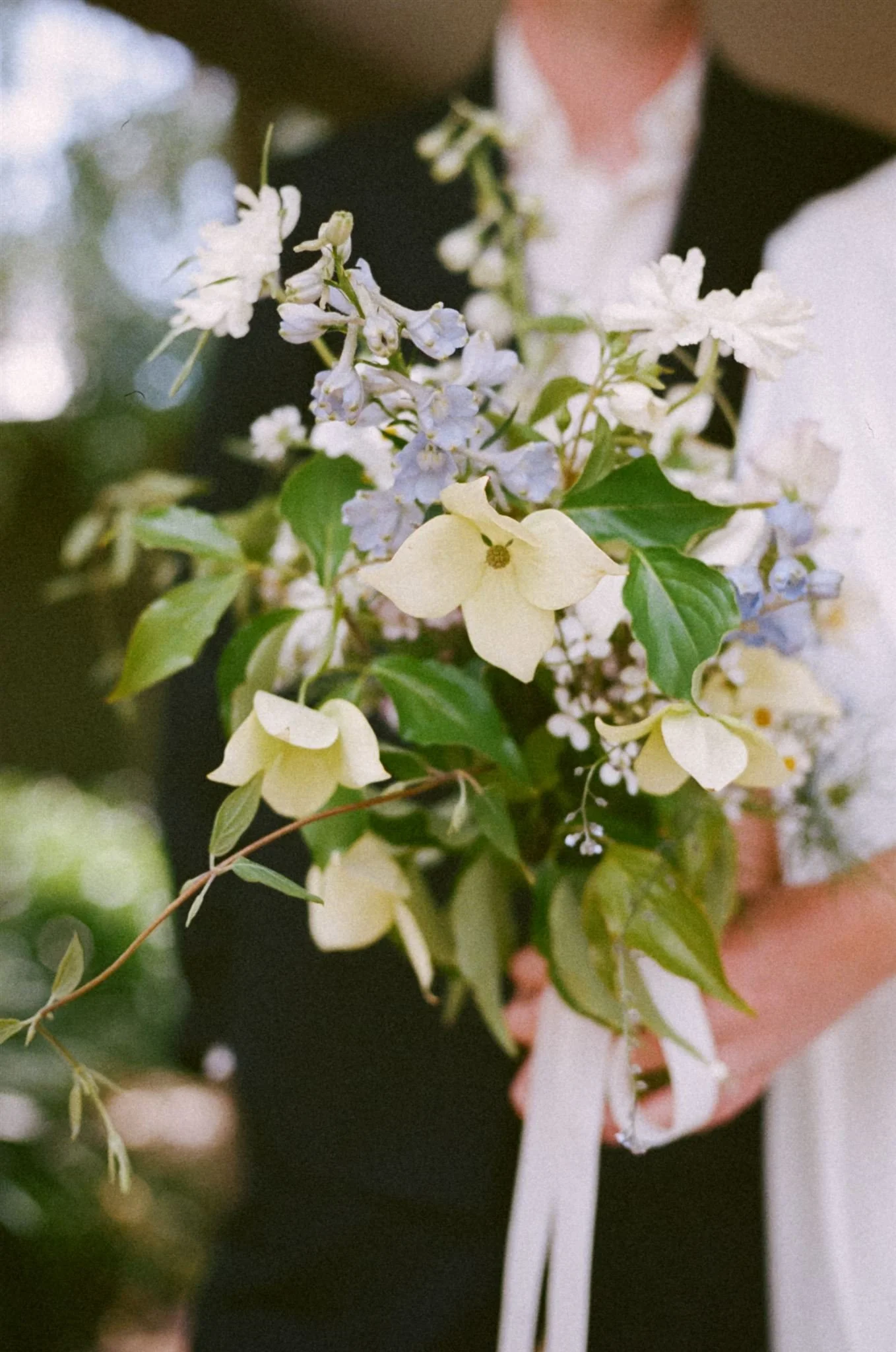 Couple holding a wedding bouquet of white and light purple flowers with green leaves.