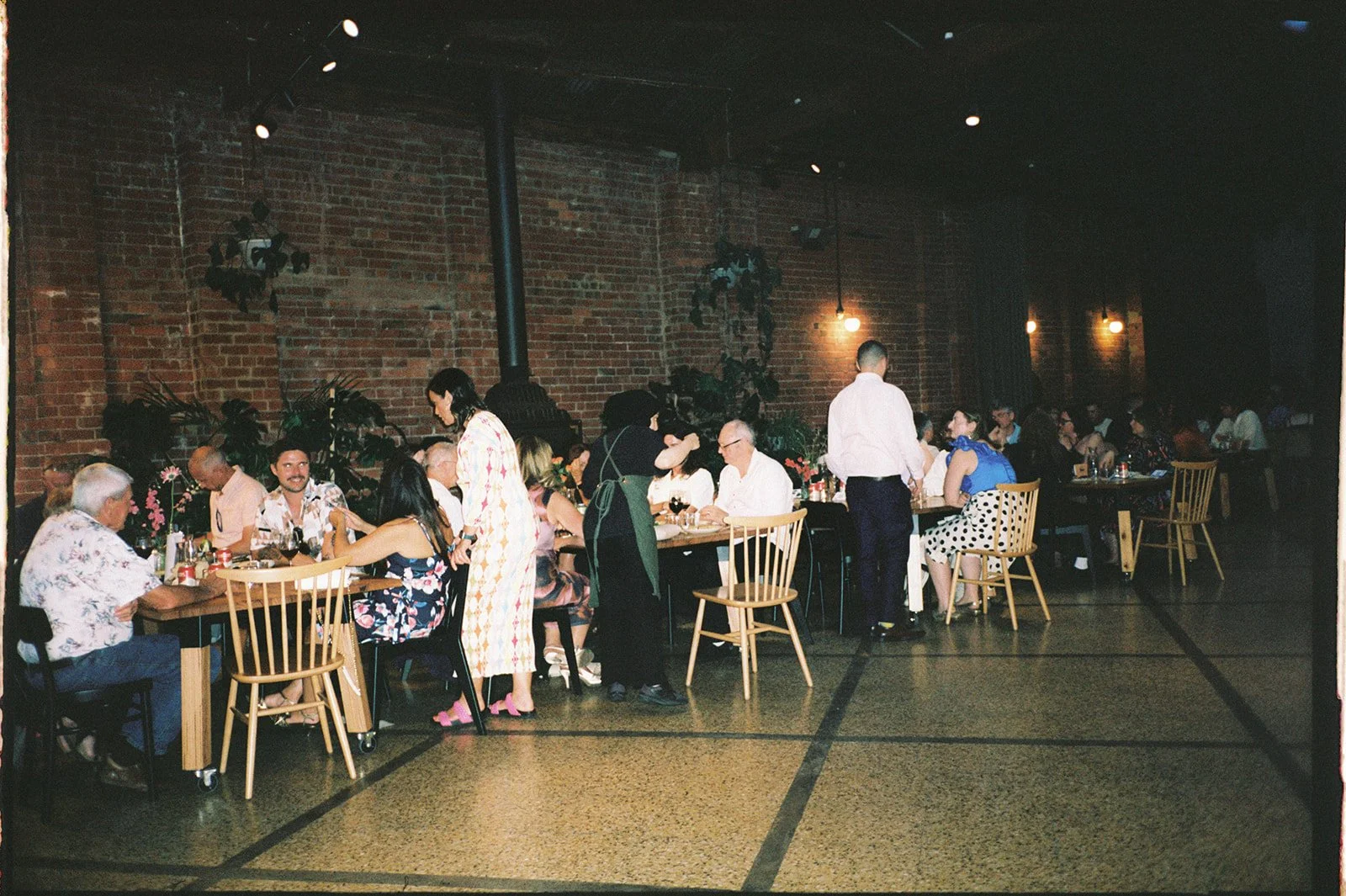 People dining at tables in a restaurant with exposed brick walls, dim lighting, and potted plants.