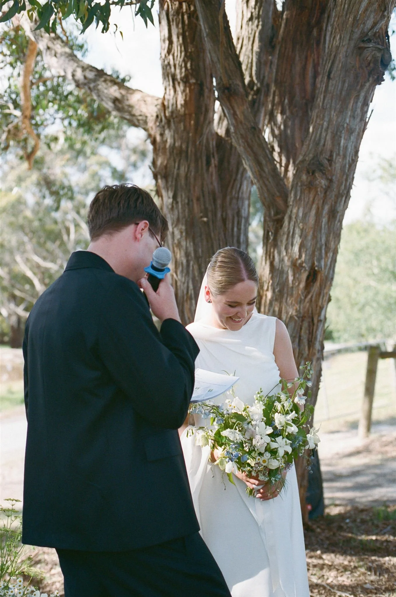A groom shares his vows with his bride at an outdoor ceremony, she smiles while holding a green, blue, and white bouquet. 