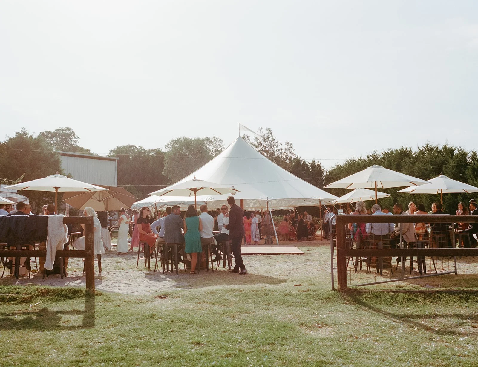 Wedding guests gather outside in colourful outfits. A wedding tent sits behind them with a white flag blowing in the wind. 