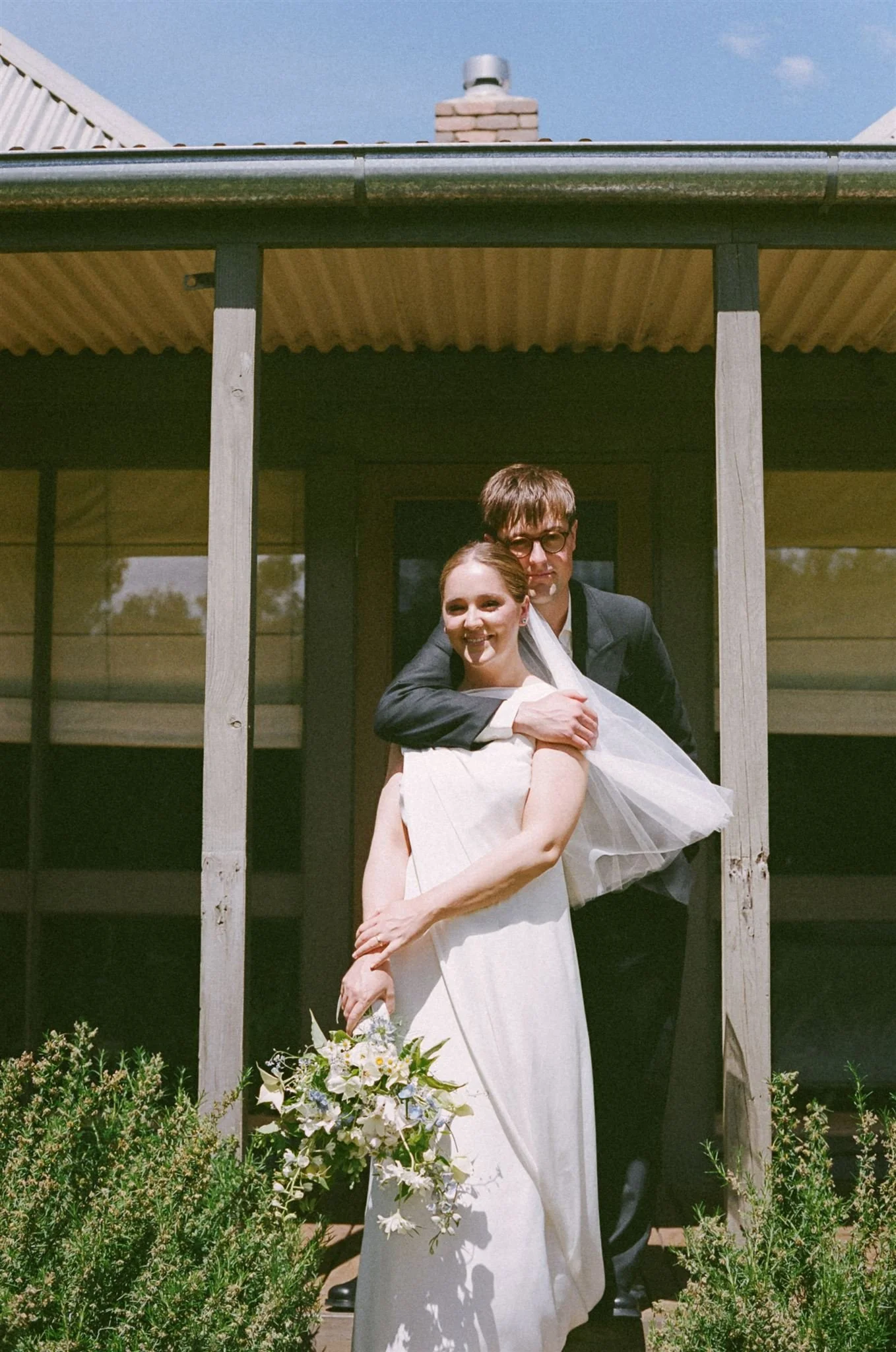 A bride and groom, standing on steps outside. The groom is hugging his bride and the bride is holding a green and white bouquet.