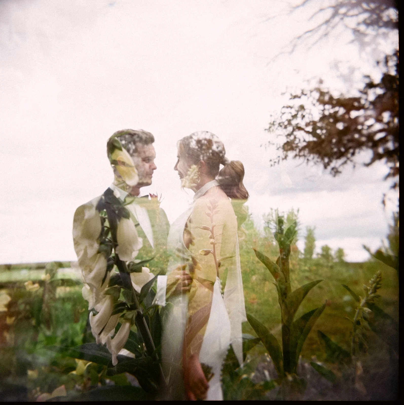 A double exposure photograph showing a bride and groom outdoors with a cloudy sky, superimposed with close-up of plants and flowers.