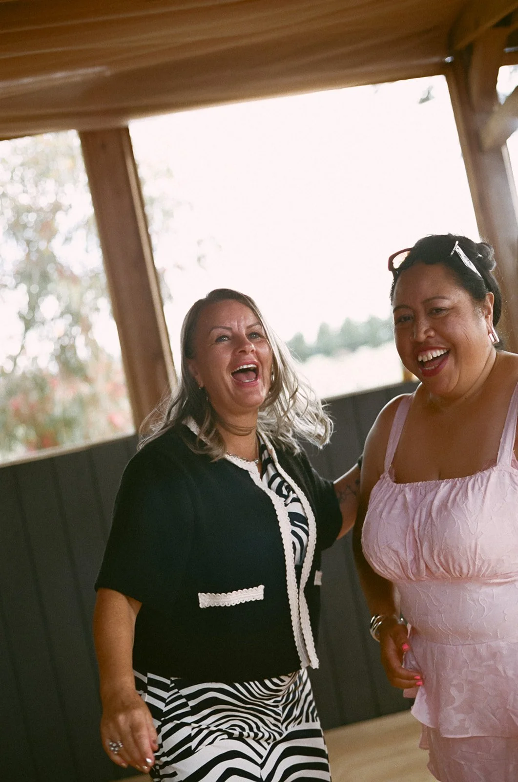 Two wedding guests dance and laugh at the reception, one is wearing a zebra stripe dress and the other is wearing a pink dress and sunglasses. 