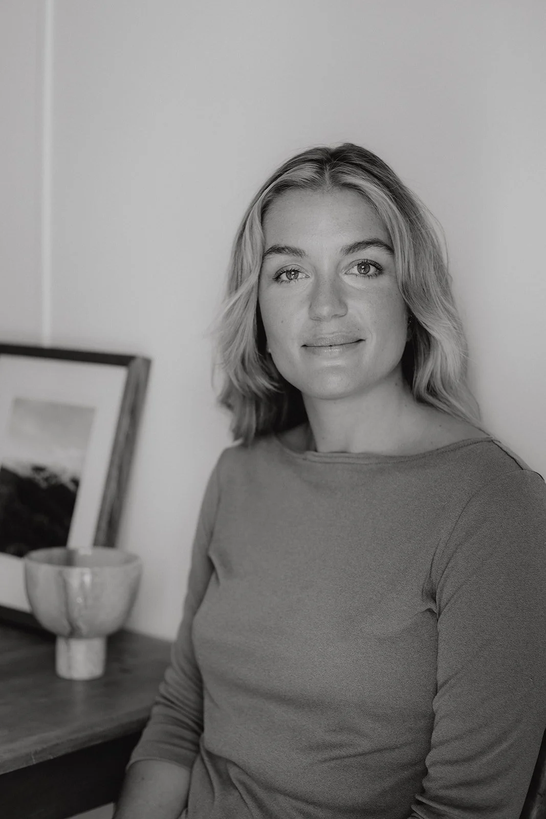 a women sits next to a table smiling at the camera, she wears a long sleeve top, the photo is in black and white.