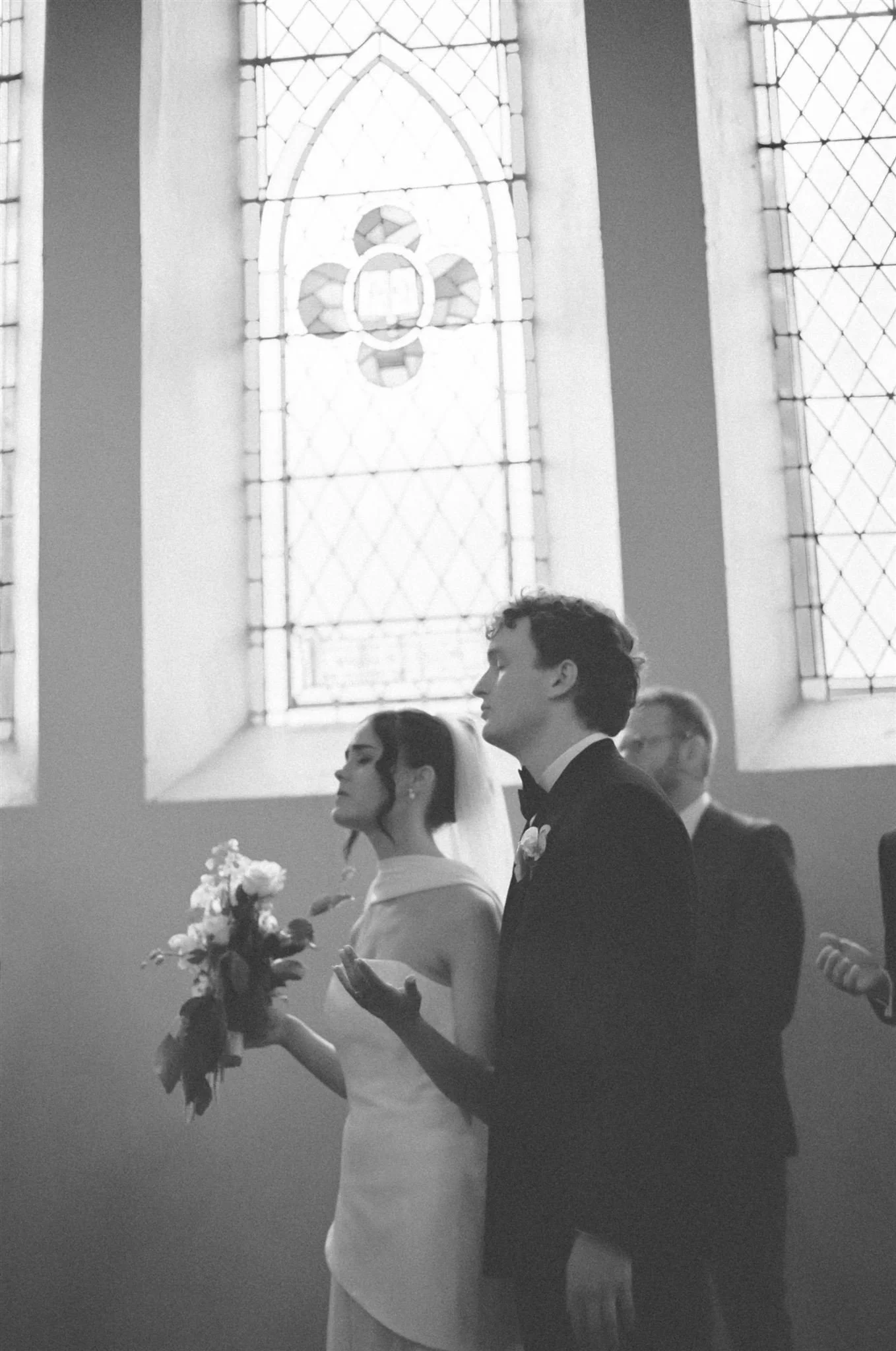 A bride and groom stand in a church during their ceremony, their eyes are closed while they listen to a hymn. The photo is in black and white.