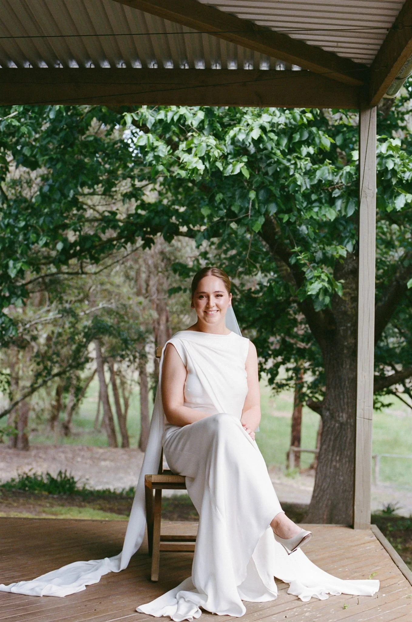A bride in a white dress sitting on a wooden chair outdoors, smiling, with a veil, on a porch surrounded by trees.