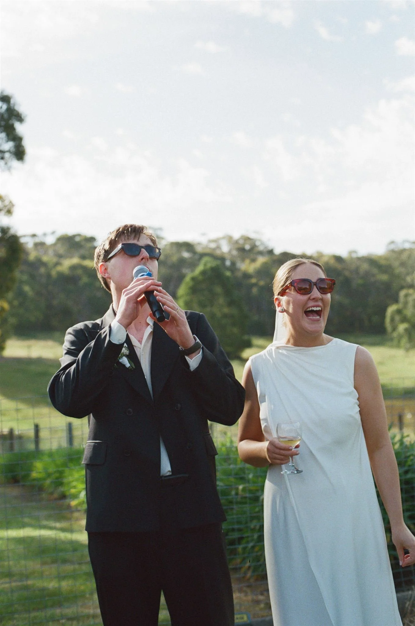A bride and groom stand in a country setting giving a speech to their guests. They are smiling and wearing sunglasses. 