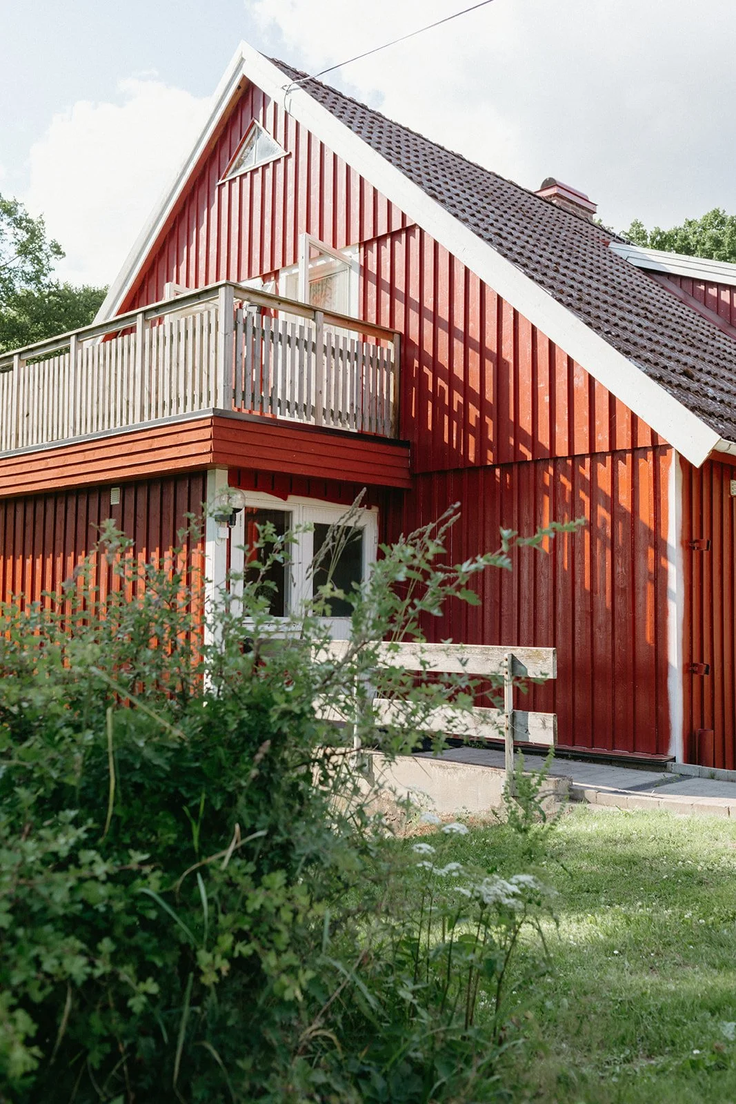 a red a frame house sits behind a green bush, the sky behind is blue with clouds.