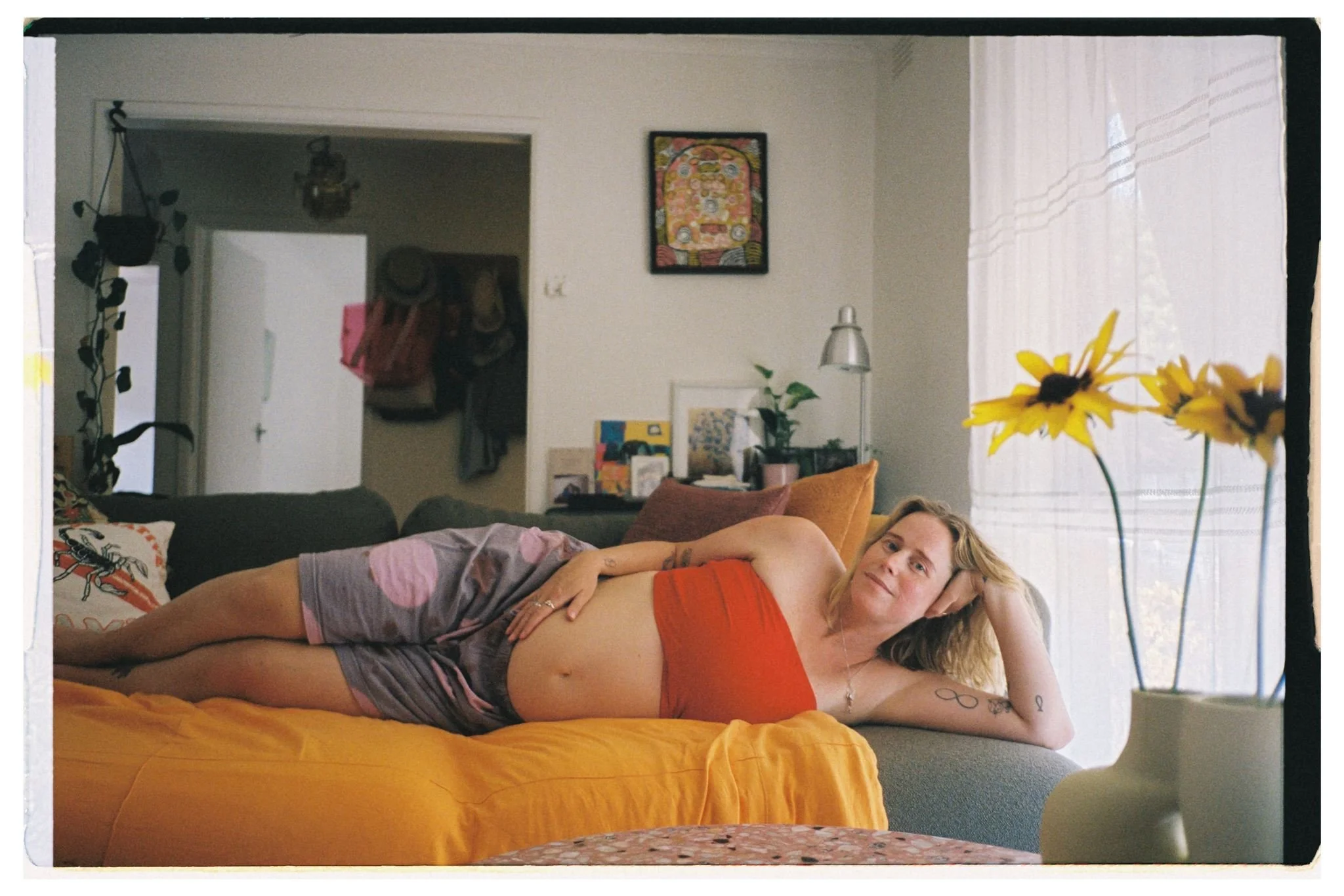 a pregnant women lays on her side on a couch holding her bump, there is a vase with sunflowers in front of her.