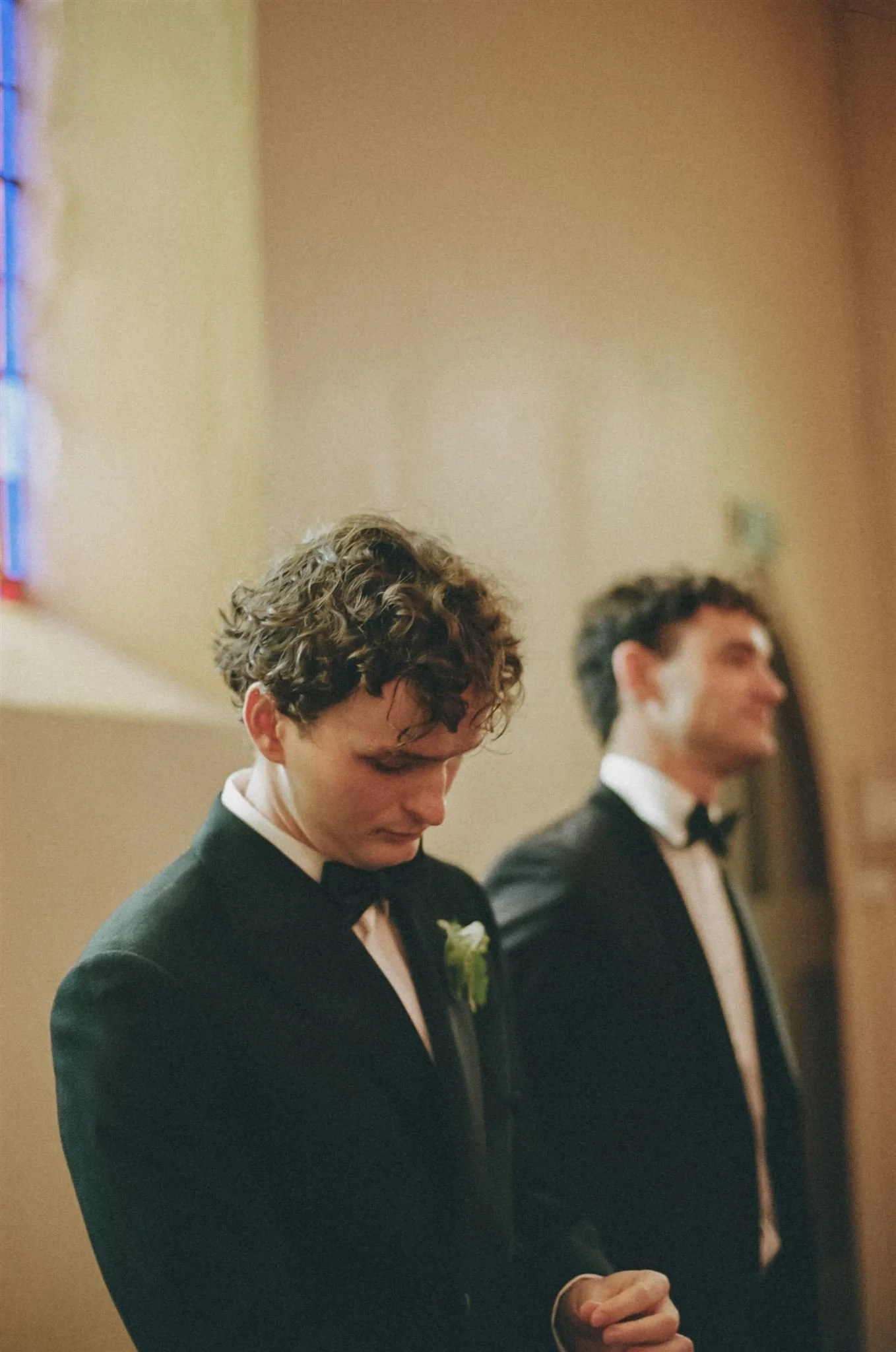 Two young men in tuxedos with boutonnières standing in a church, head bowed in prayer or reflection during a wedding ceremony.