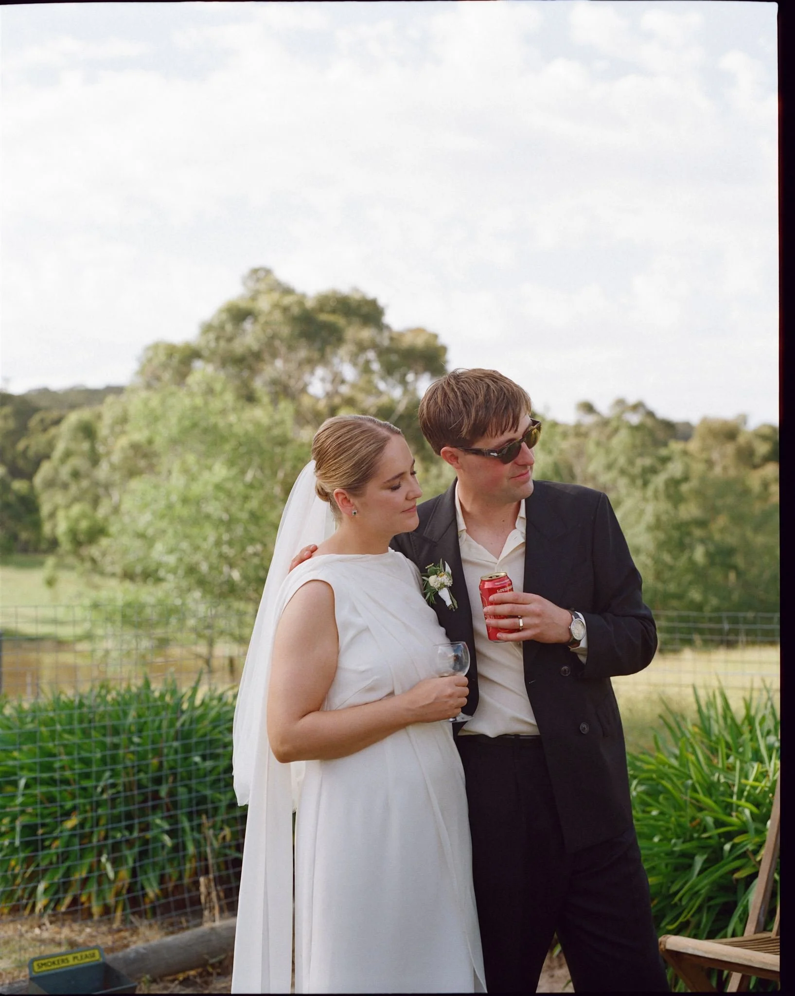 A bride and groom stand at an outdoor reception listening to a guest give a speech. The groom has his arm around the bride and holds a beer. 