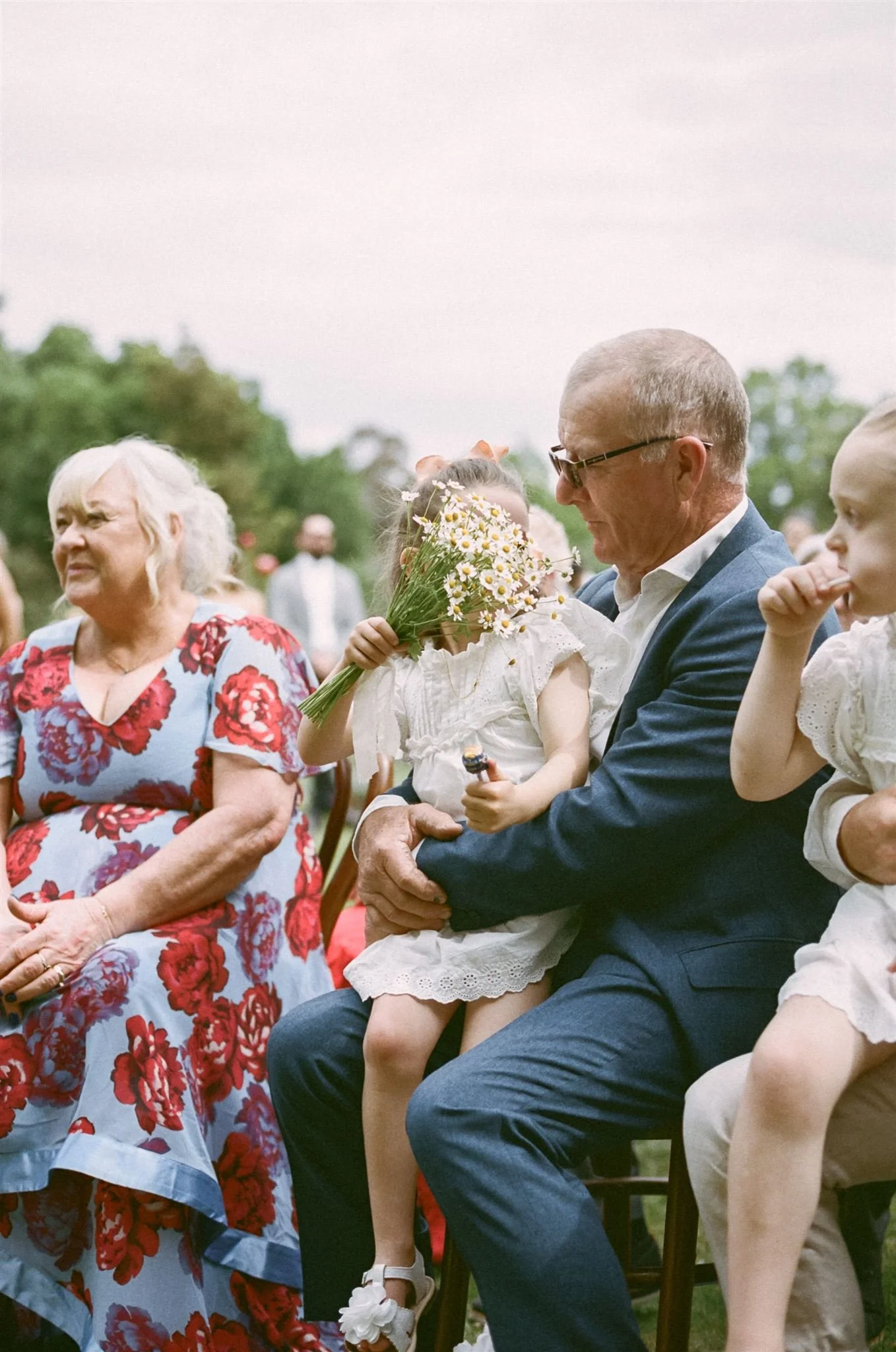 The bride and grooms parents are sitting at the front of the ceremony holding the flower girls, the girls are waving a bunch of daisies. 