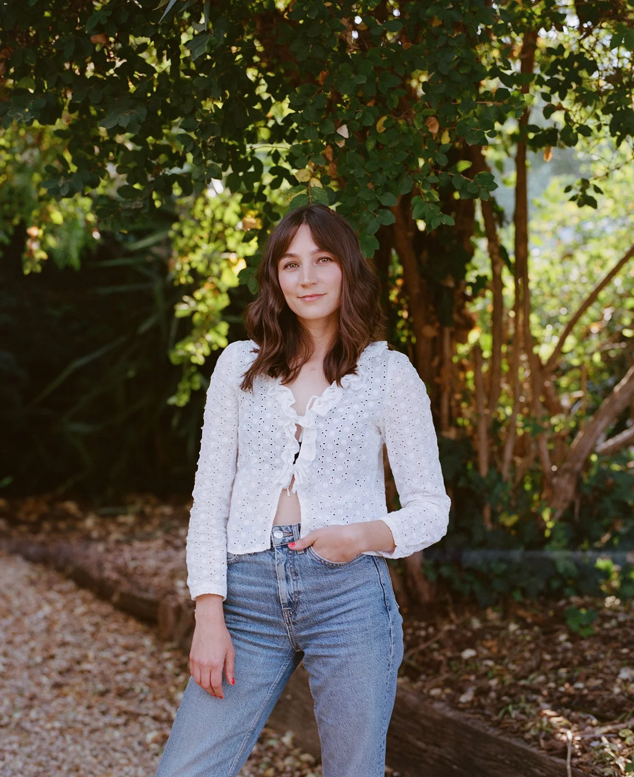 A film photograph of a women standing in a garden with her hand in her jeans pocket, the sun is shining through the tree leaves.