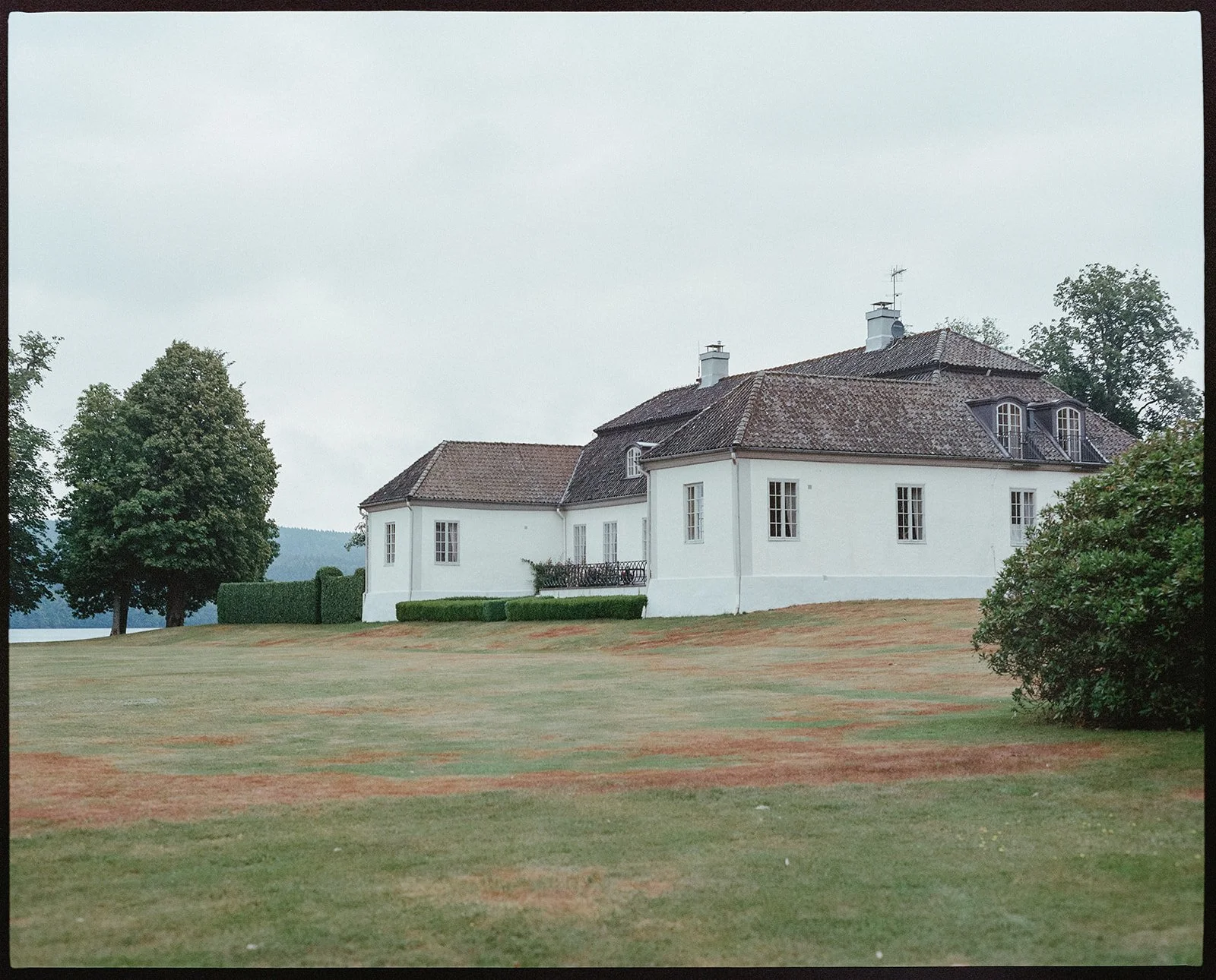 A large white manor house sits in a green field with a lake in the background. 