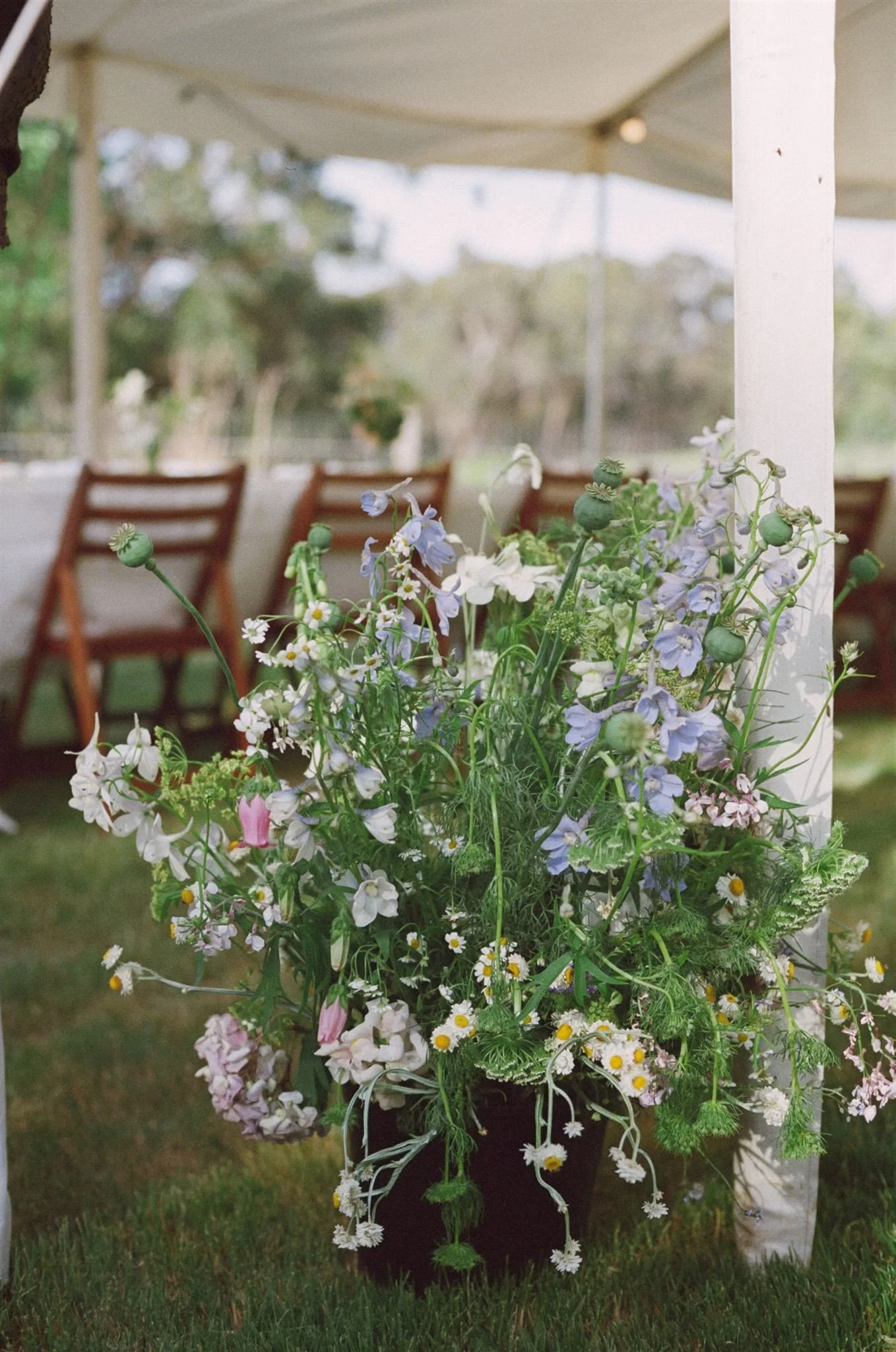 A bucket of wedding flowers sits under a wedding tent. The flowers are green, white, blue, and pink. They look like wild flowers.