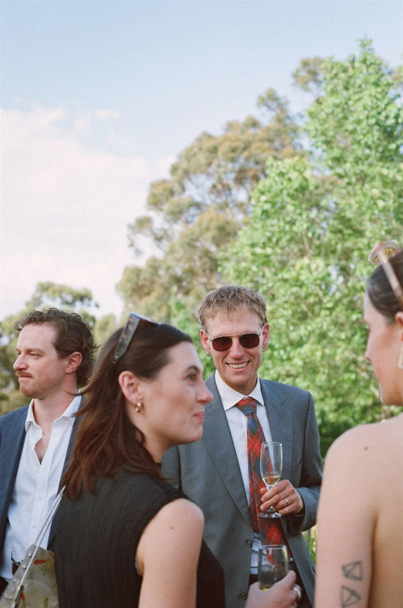 Wedding guests mingle outside during the reception. They are holding glasses of champagne and tree's are behind them. 