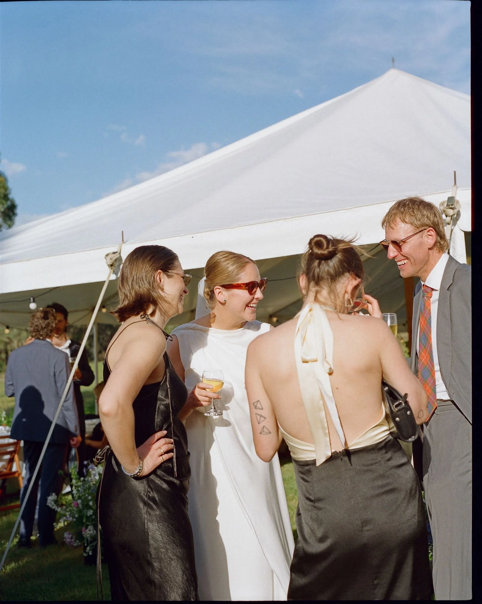 A bride stands with her wedding guests, they are laughing and the sun is shining. You can see the white wedding tent behind them.