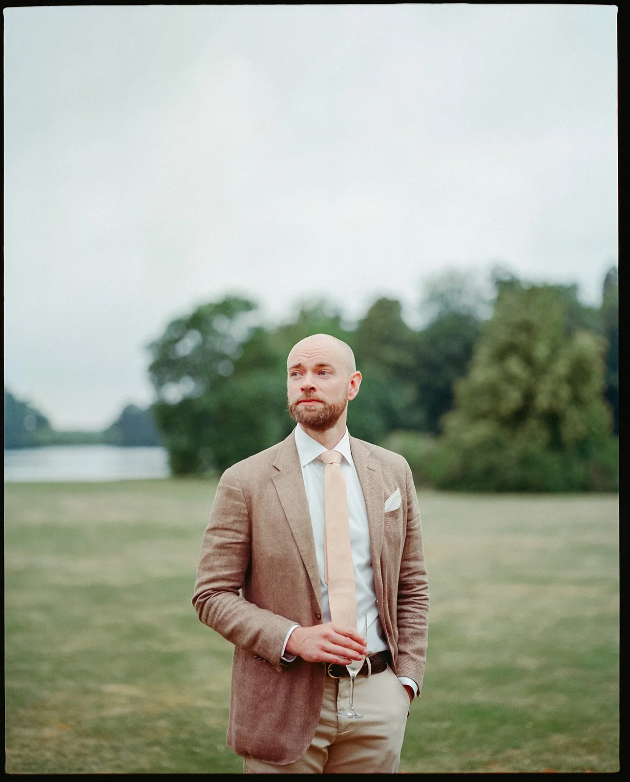 A groom stands in the middle of the field in a brown jacket, he looks off to the distance. The photo is taken on medium format film.