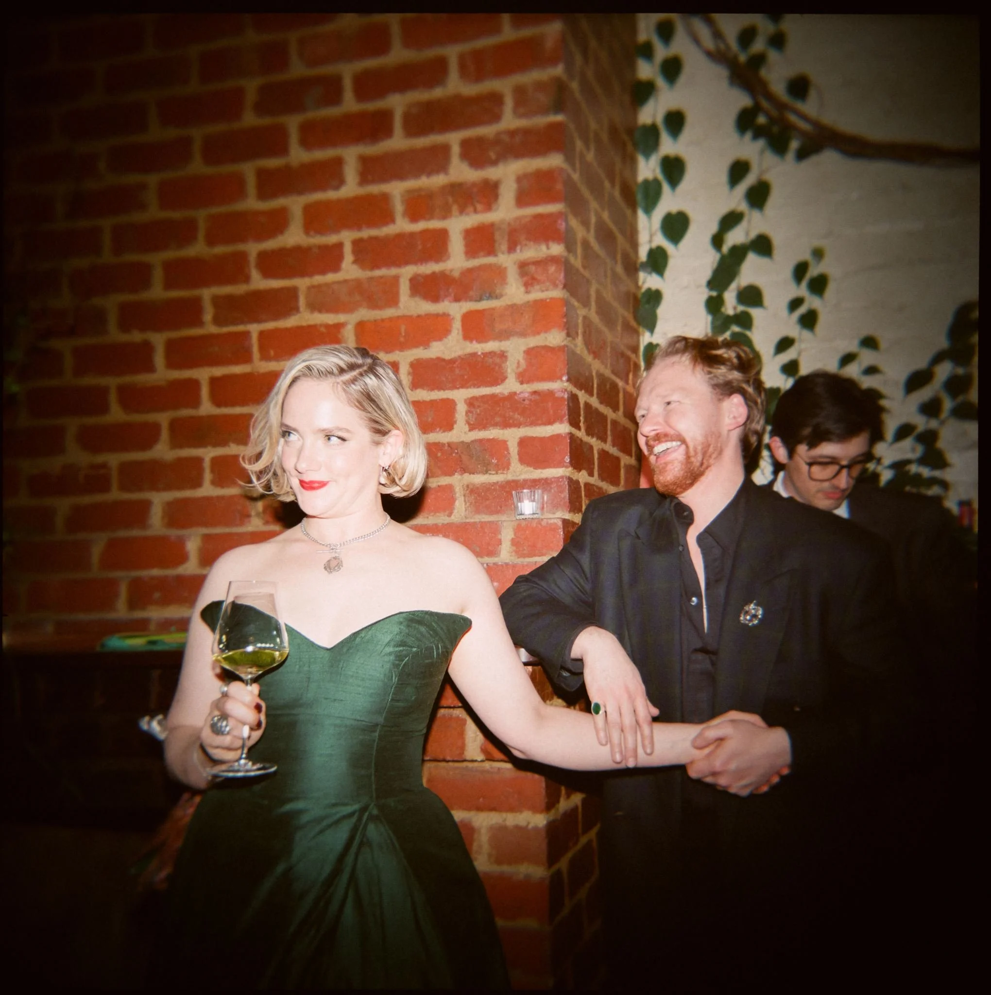 Annabelle and Dom sharing a relaxed moment against a red brick wall at Old Palm Liquor, with Annabelle wearing her bespoke green wedding dress.