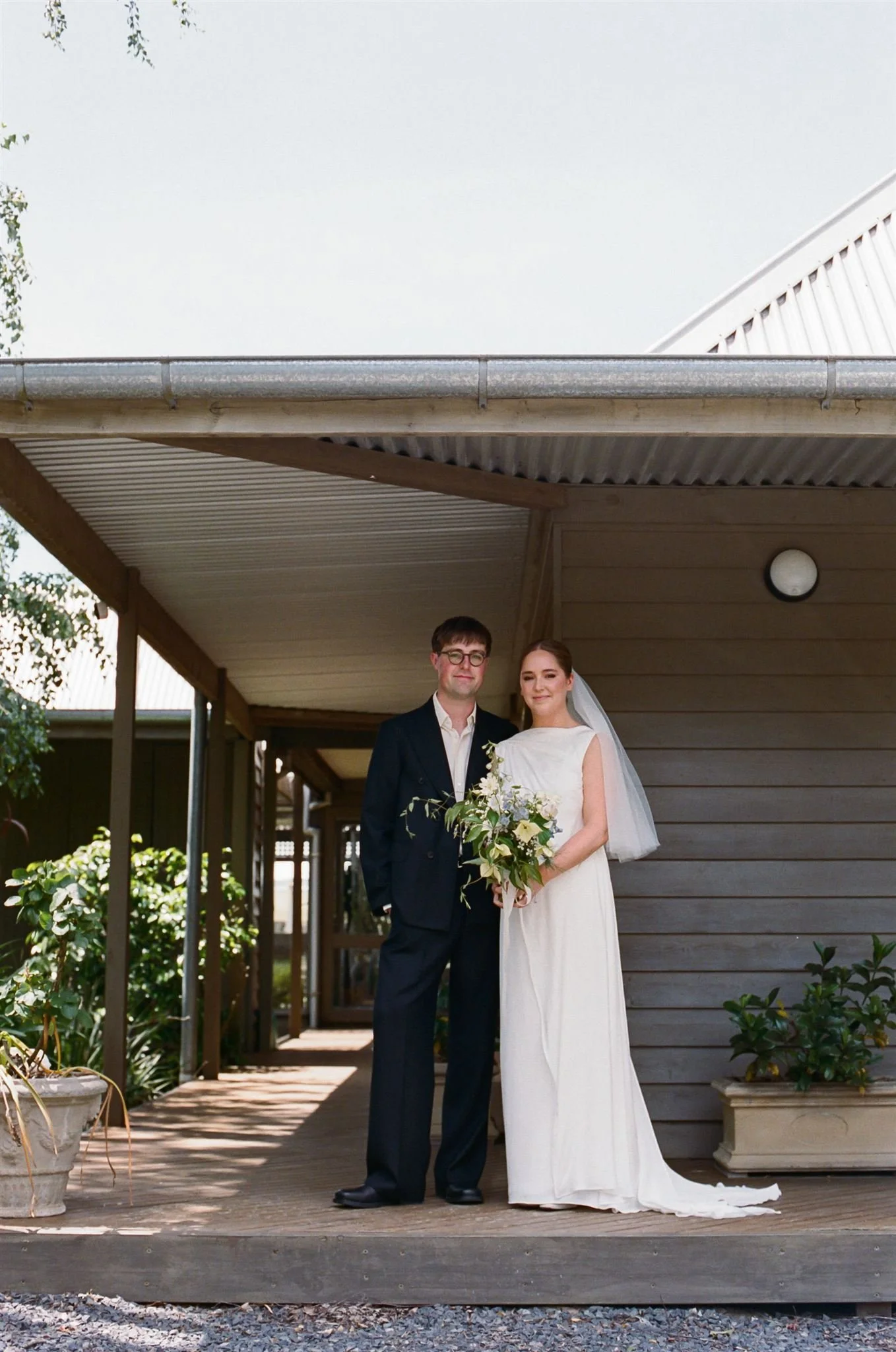 A bride and groom standing together outside on a wooden porch, dressed in wedding attire, with the bride holding a bouquet of flowers.