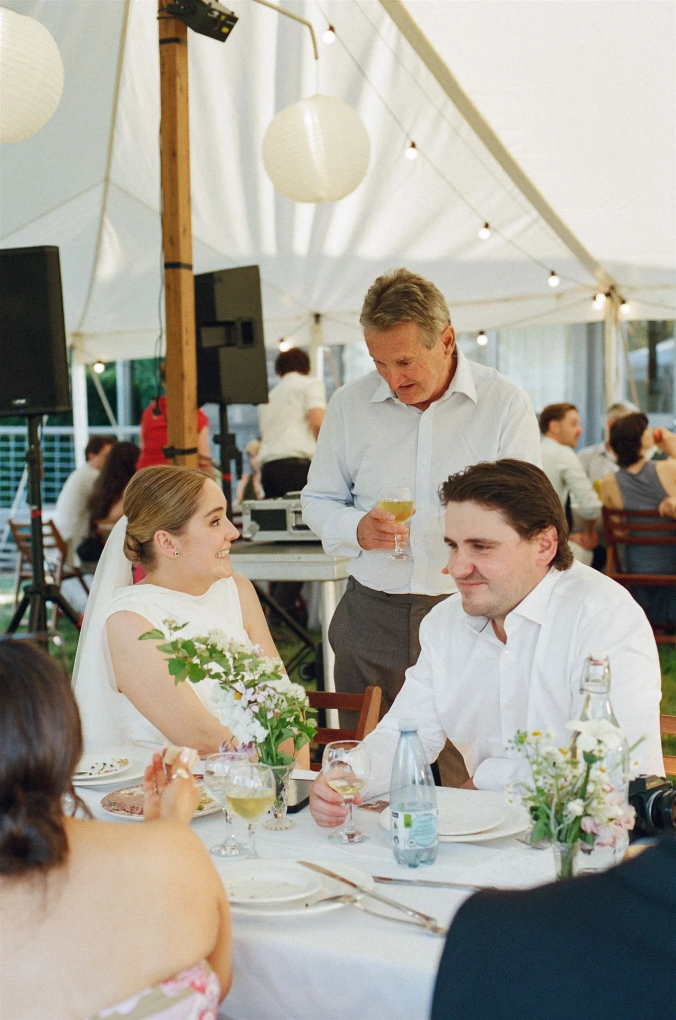 A bride sits at the wedding reception table next to her brother, her dad stands next to her as they talk. There are green flowers on the table.