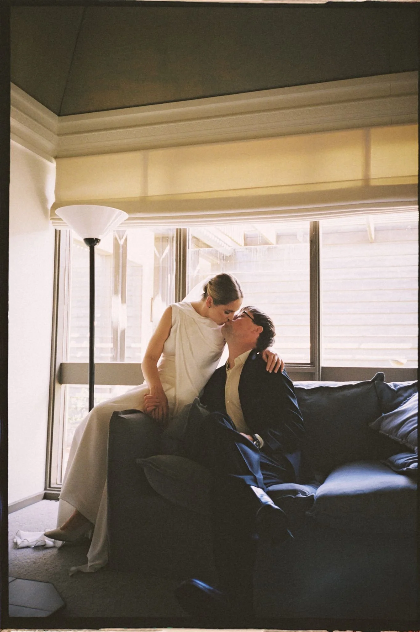 A bride in a white wedding dress and a groom in a dark suit sitting on a black couch, sharing a kiss in a bright room with large windows and natural light.