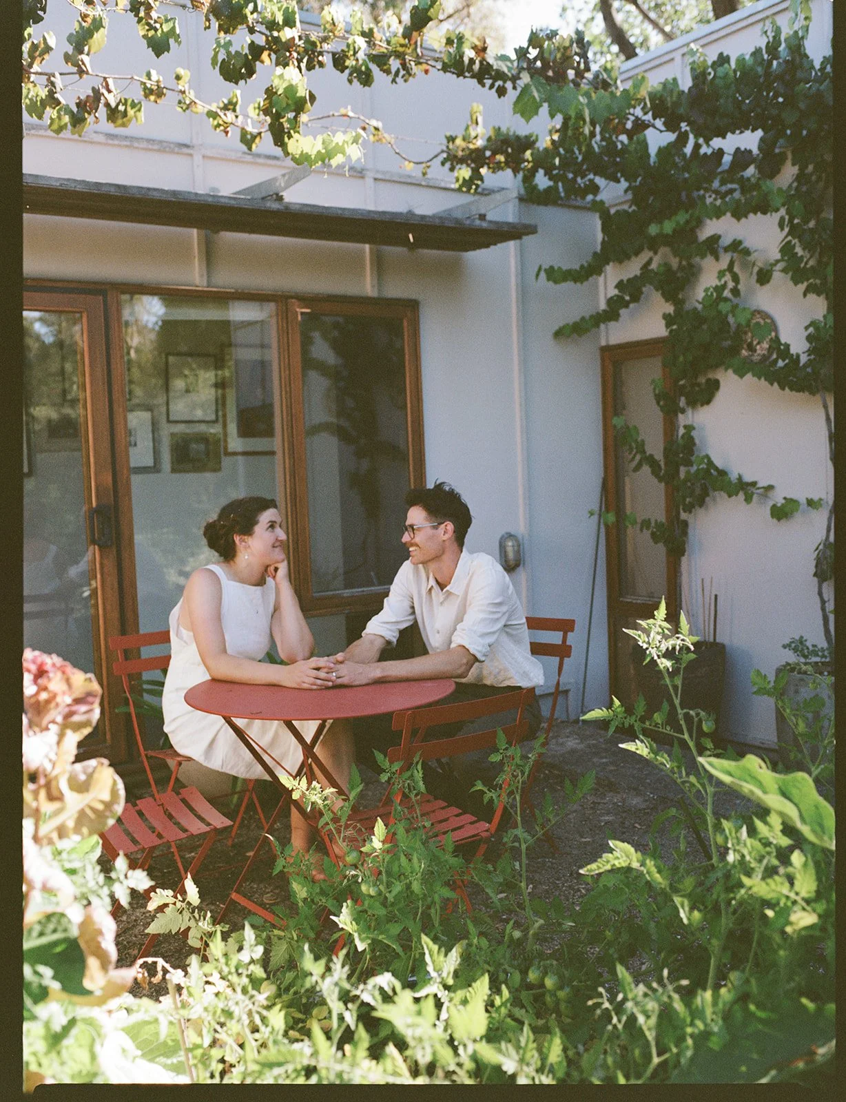 A bride and groom sit in the garden around a little red table holding hands. There are tomato plants around them.