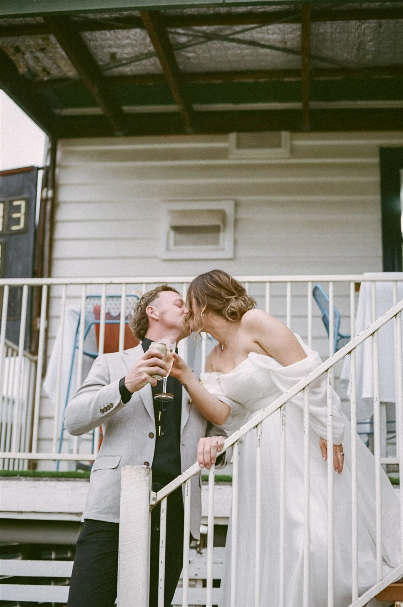 A bride and groom standing on the steps of their reception venue. They are holding champagne and sharing a kiss.