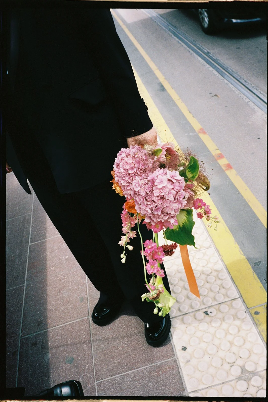 A bride is standing waiting for a tram, she is wearing a black suit and holds a bouquet of pink, orange, and green flowers. 
