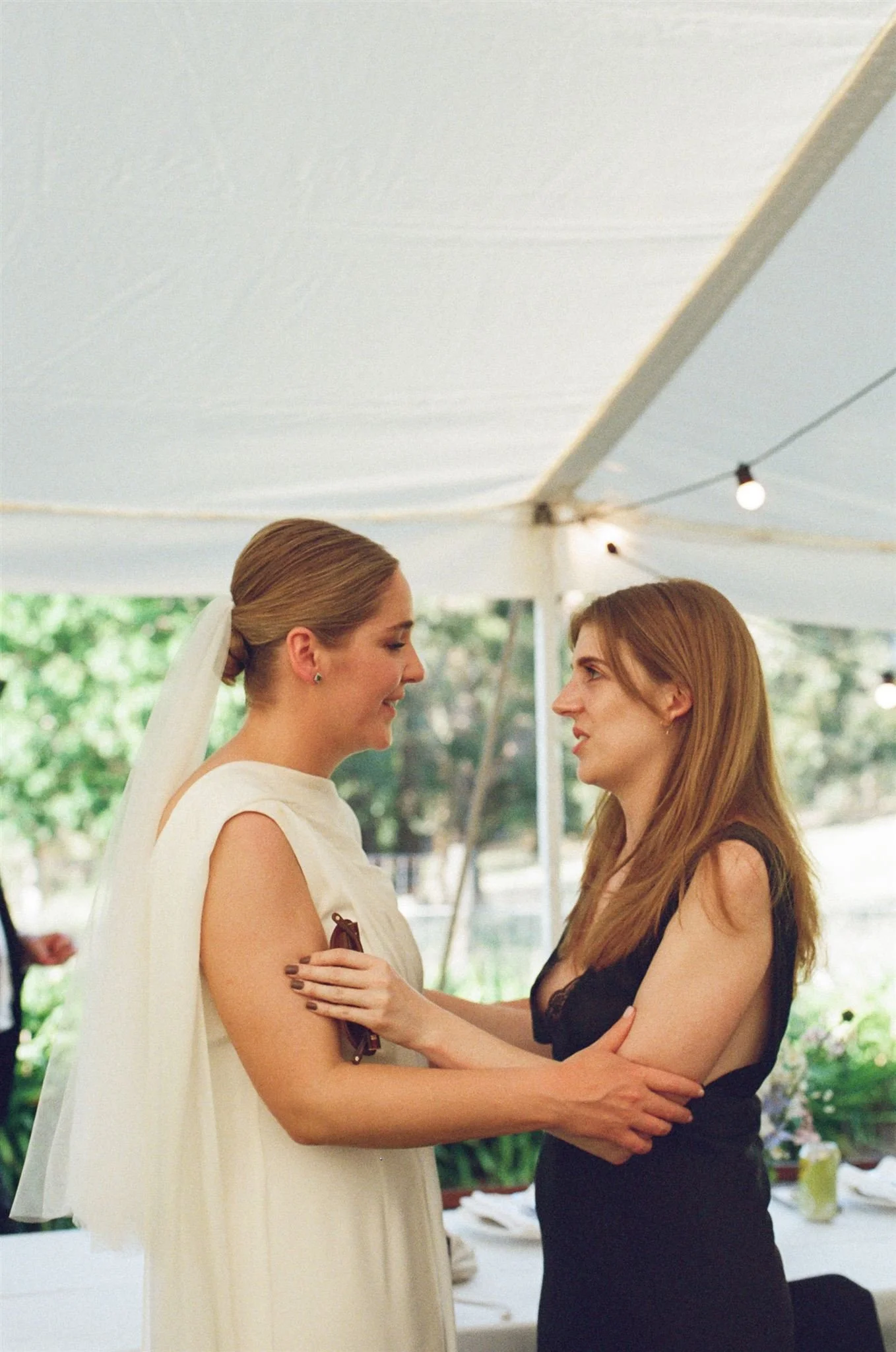 A bride and wedding guest stand together, they are talking and holding each others arms. 