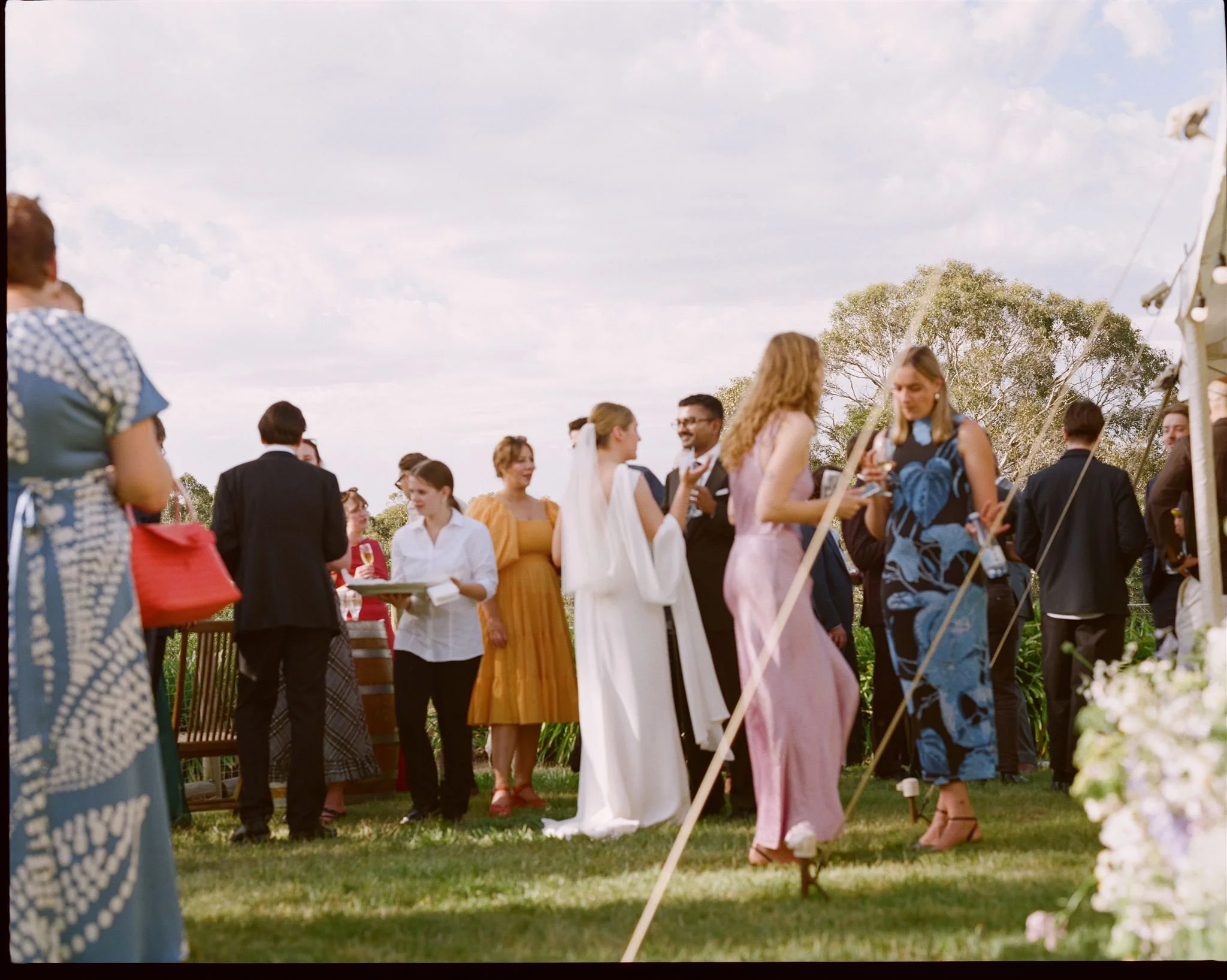 Wedding guests gather outside at the reception, the bride is in the middle of the guests and the sky is blue with light clouds. 