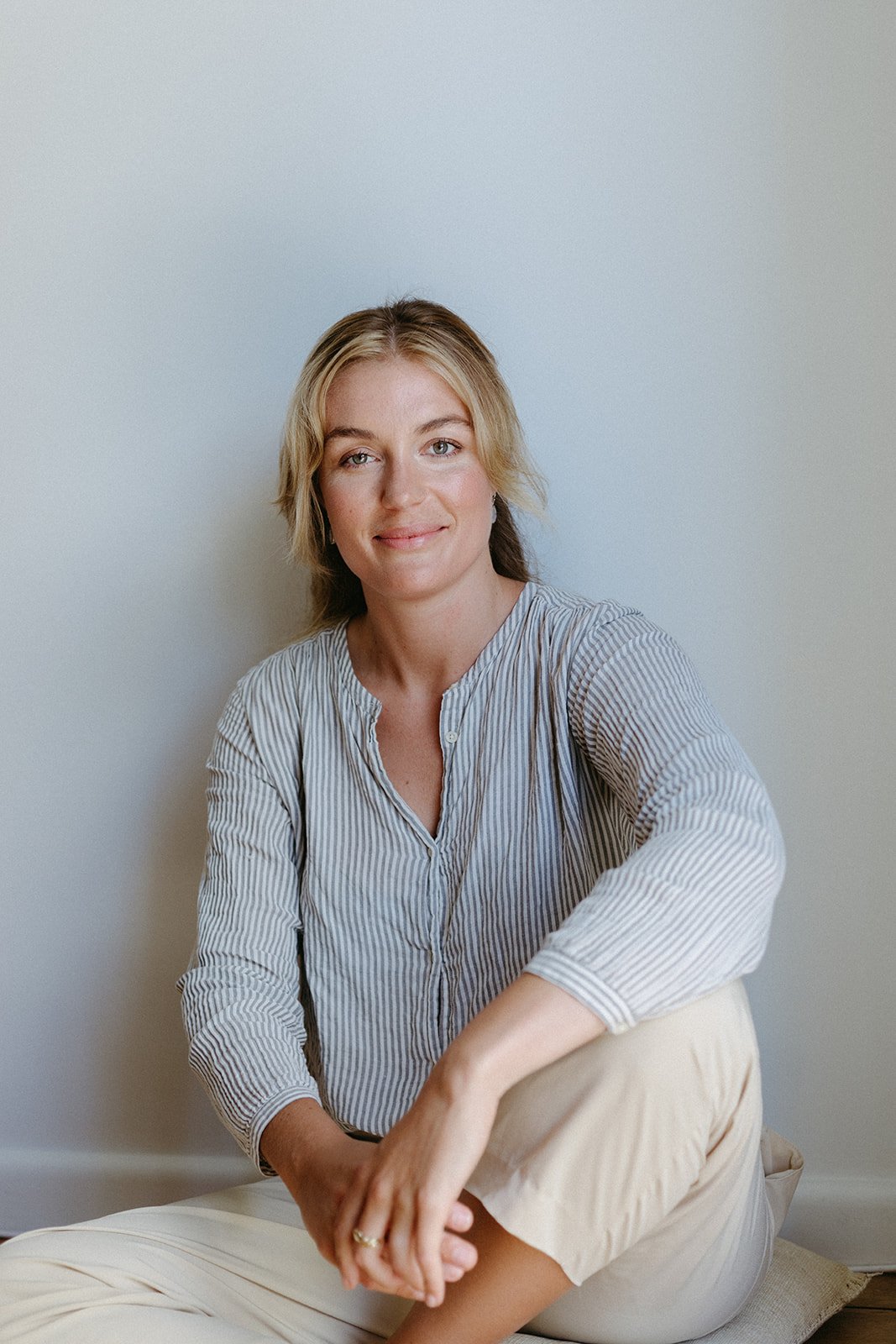 A women sits against a blank wall smiling at the camera, she is sat in window light resting an arm on a knee.