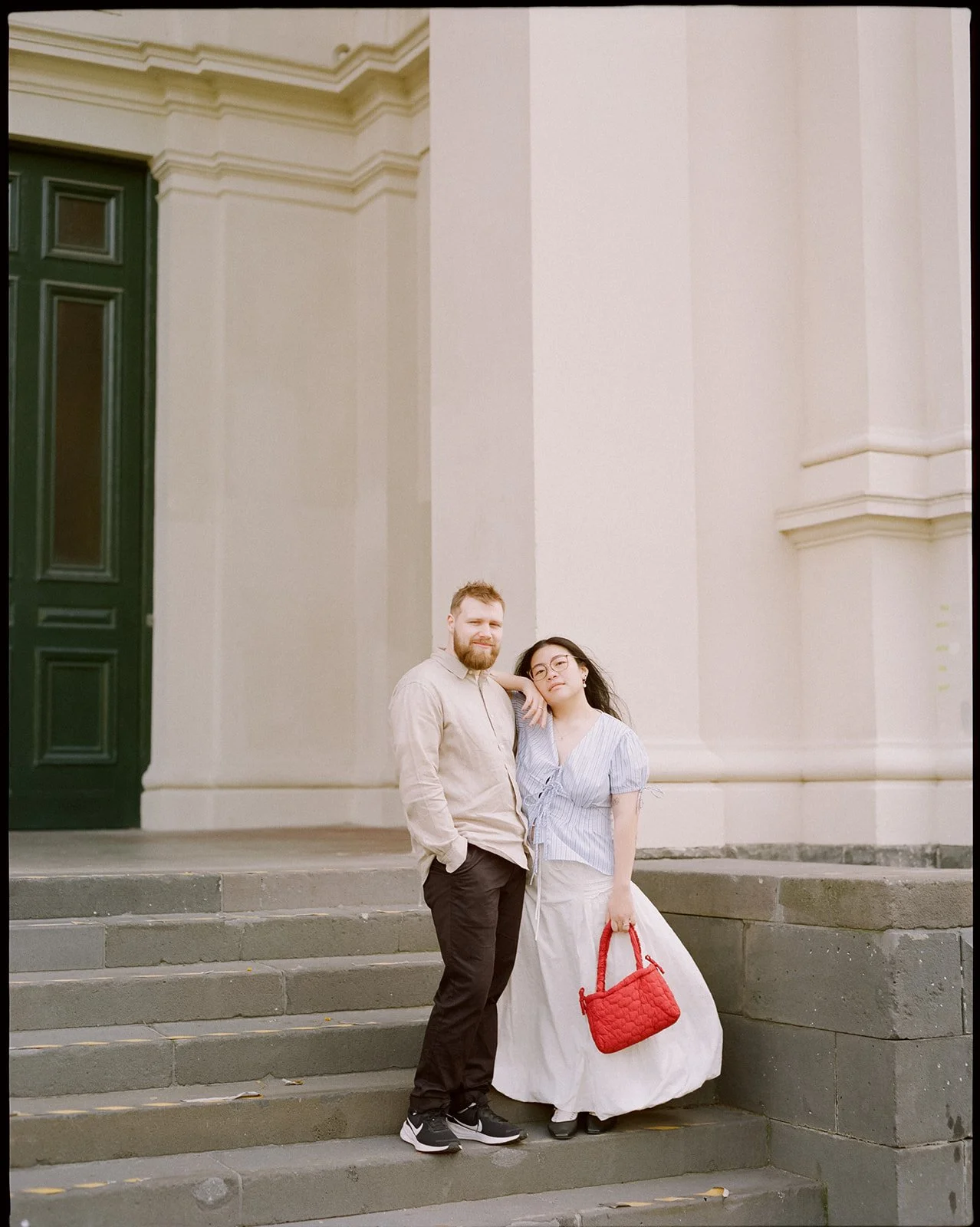an engaged couple stand on some steps together, the women rests her arm on her fiancé's shoulder, her skirt blows in the wind.