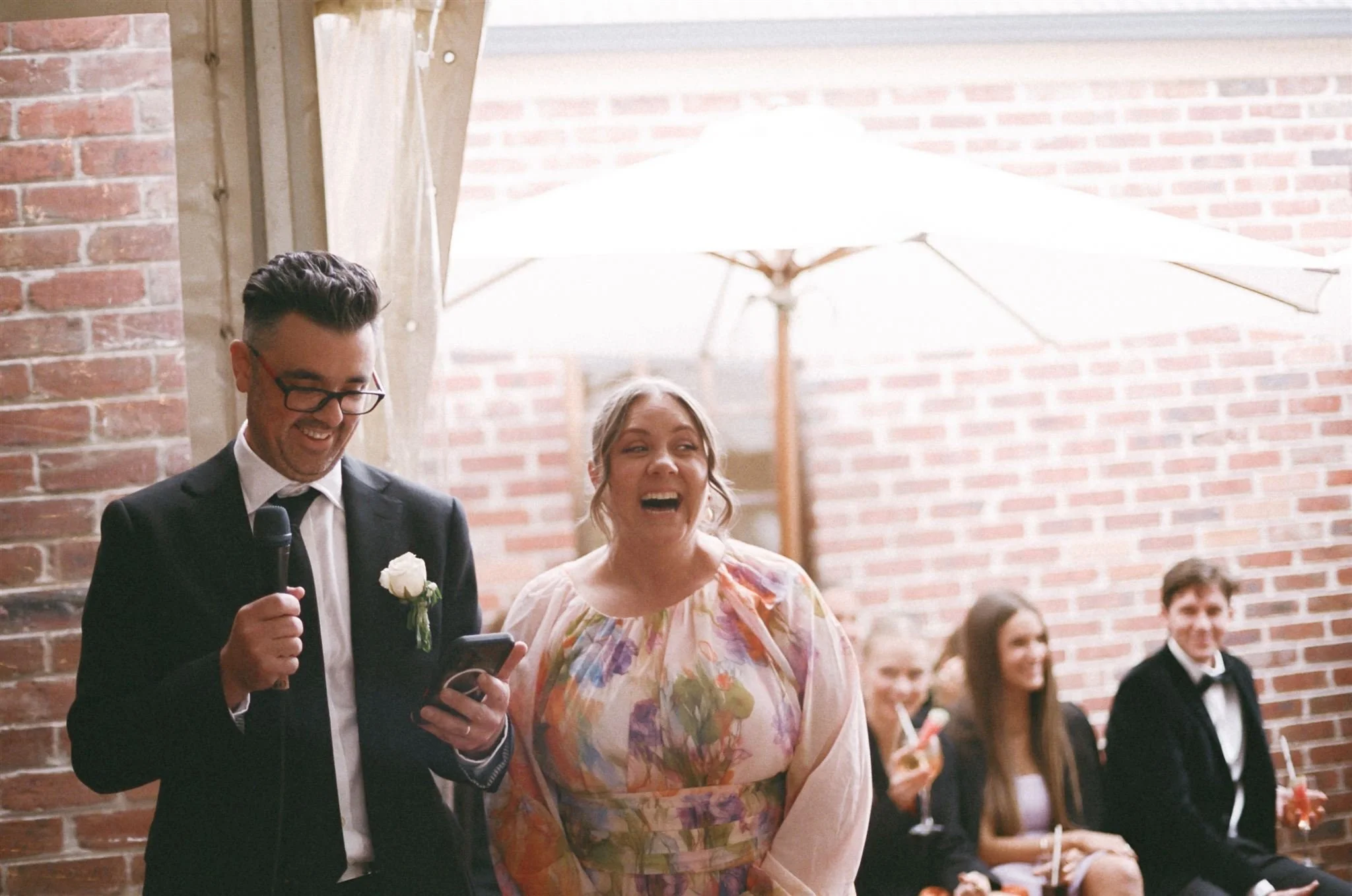 A man in a suit with glasses holding a microphone and reading from his phone, standing next to a woman in a floral dress laughing at a wedding reception. In the background, guests sit near a brick wall under a large white umbrella.