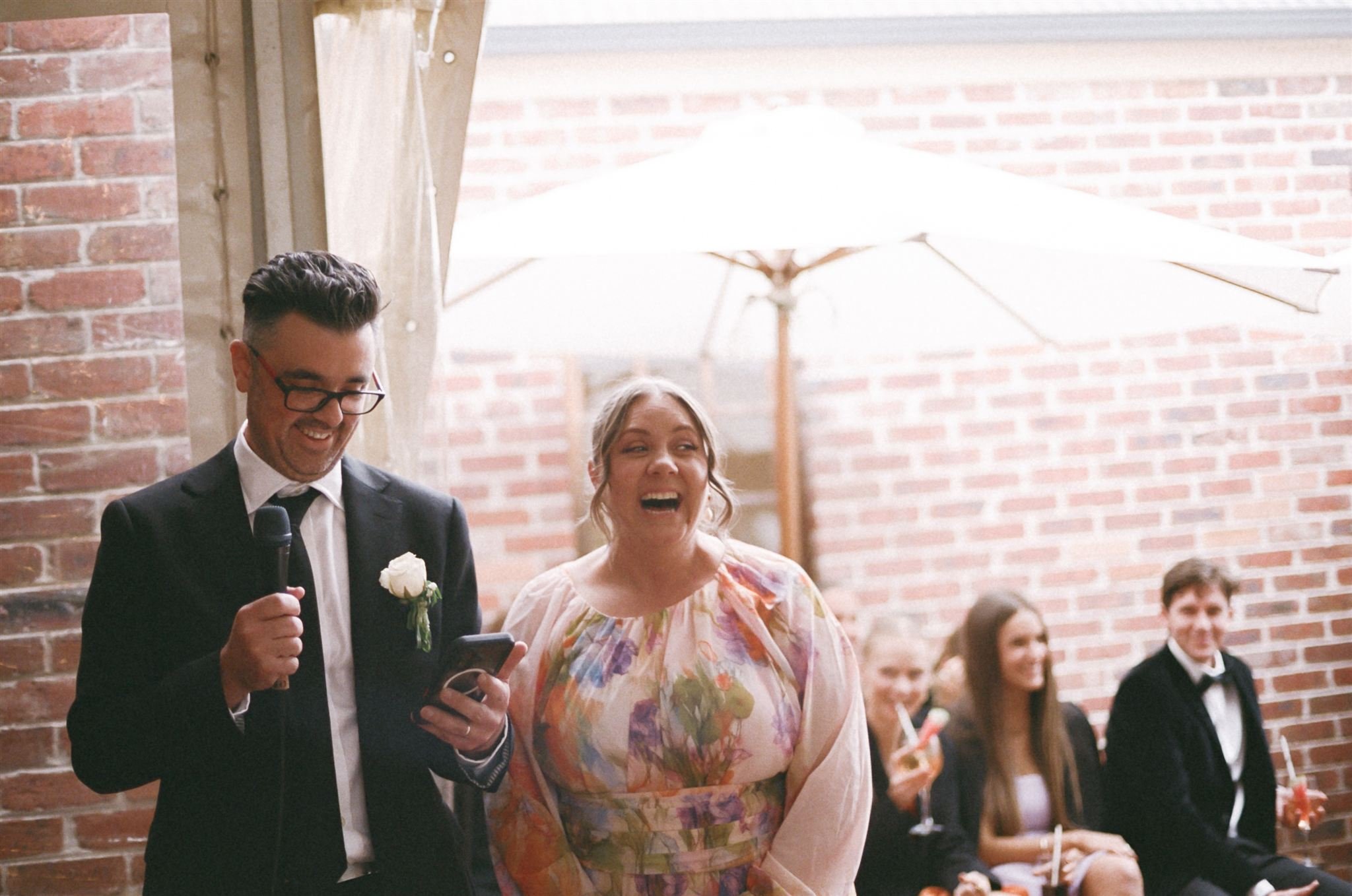 a mother and father in law give a speech at their sons wedding, they are stood infant of a brick facade, the grooms mums is wearing a floral dress.