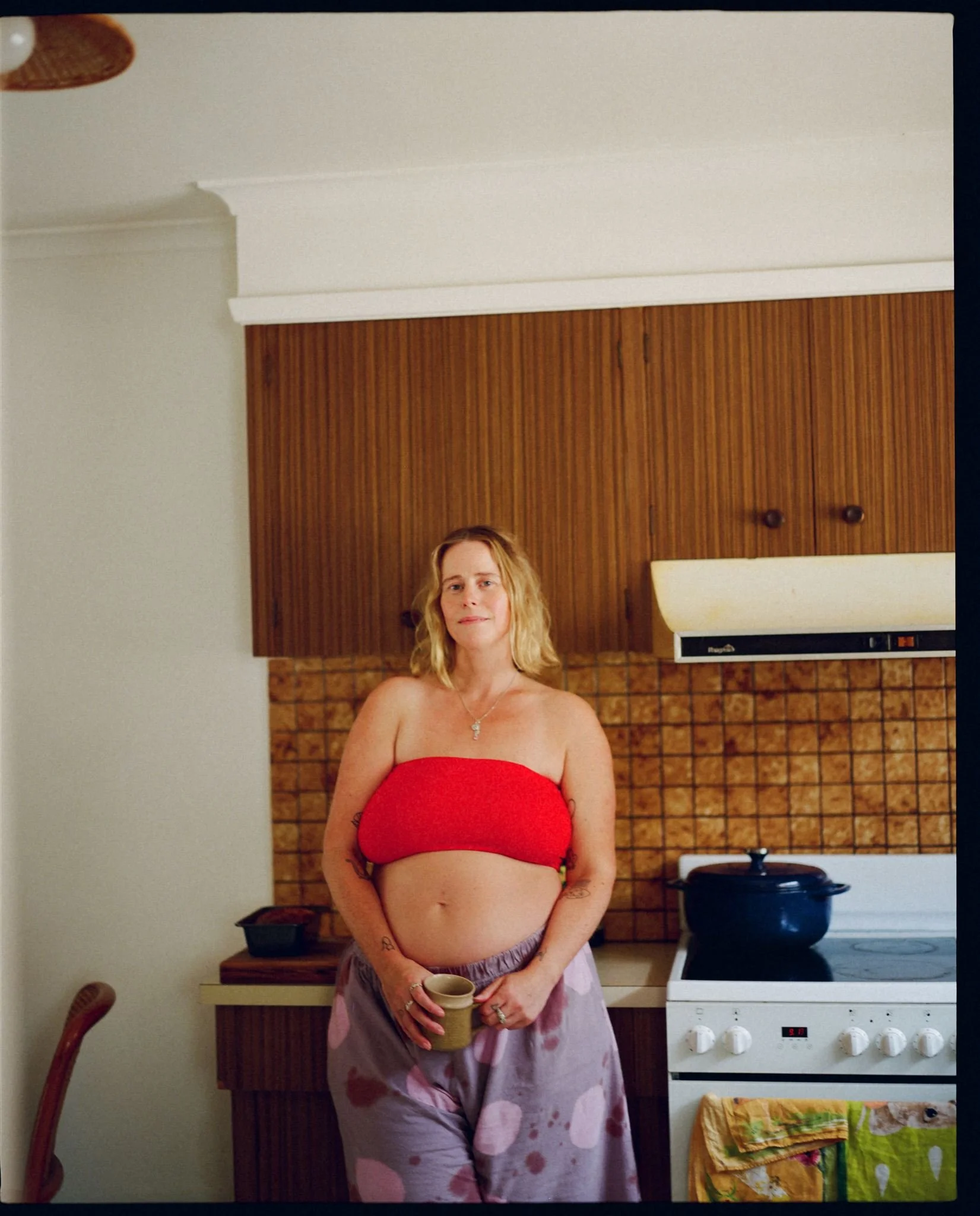 A pregnant lady stands in her kitchen, she is holding a mug in her hands and is wearing a red boob tube and purple shorts.