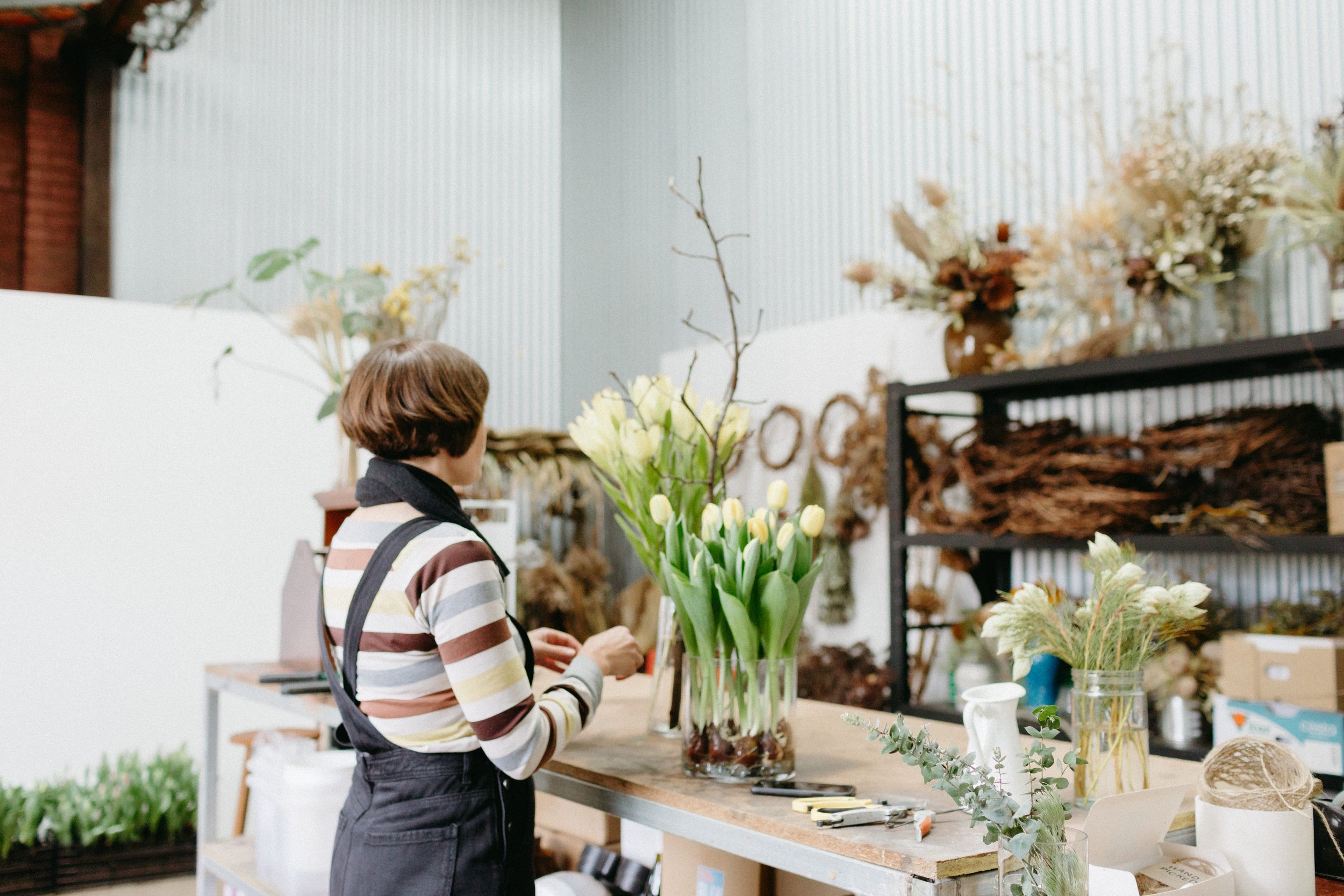 a women is standing in her workshop arranging tulips, she is wearing dark dungarees and a striped top.