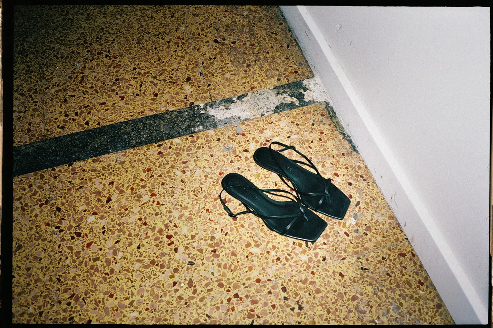 A wedding guests shoes sit next to the dance floor, they are black and scrappy. The photo was taken with flash.